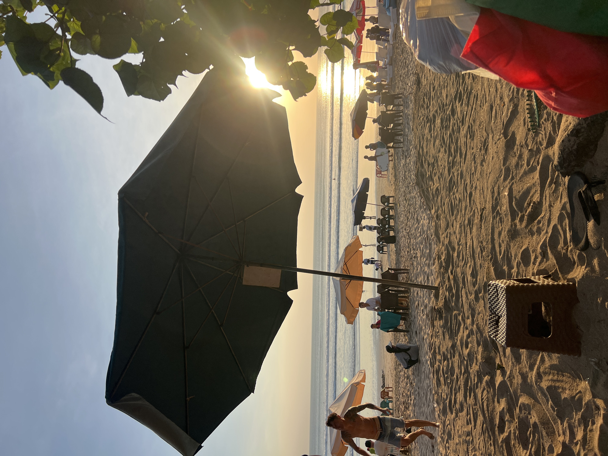 The image depicts a beach scene in Kuta, Indonesia, at sunset. The foreground is dominated by sand dunes and shadows cast by a large, dark umbrella. A small brown plastic stool sits on the sand in the mid-ground. In the background, the ocean stretches out to the horizon, with gentle waves breaking on the shore. Several beach umbrellas, both open and closed, are scattered across the sandy area. Groups of people are visible, some sitting at tables under umbrellas, while others are standing or walking. A man with tattoos is walking in the left mid-ground, and another person is sitting on the sand nearby. The setting sun casts a warm glow over the scene, creating a golden hue on the water and sand. Lush green leaves and branches of a tree are silhouetted against the bright sky in the upper right corner. The atmosphere is relaxed and suggests a popular tourist spot during evening hours.