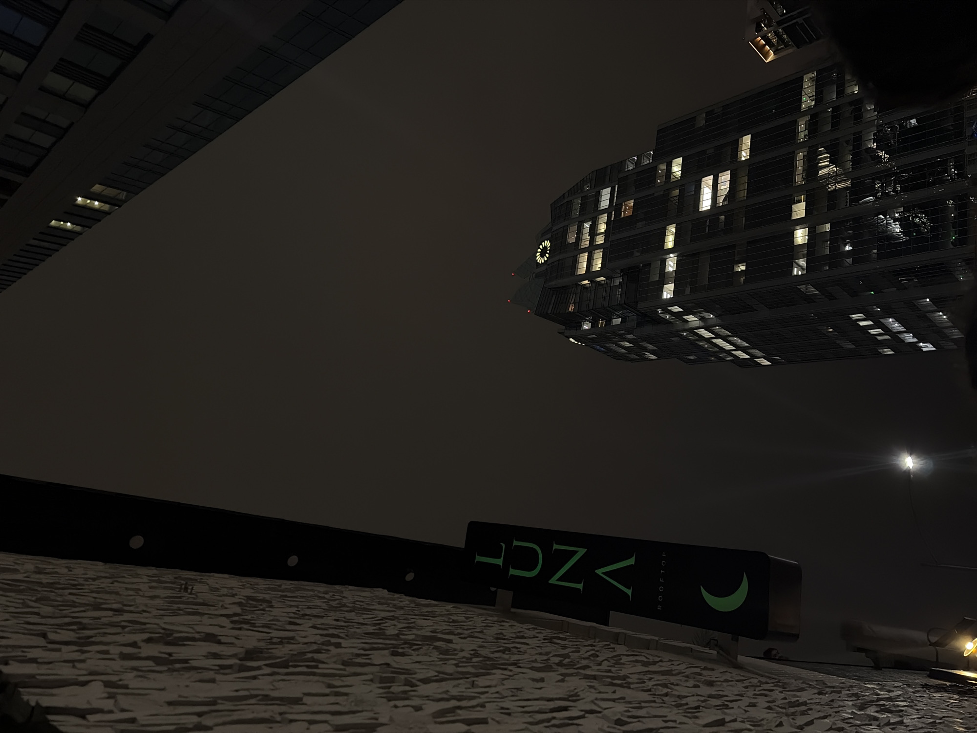 The image, taken at night, shows a low-angle perspective of a building in Austin, United States of America. The focus is on the rooftop with a sign that reads "VNZA ROOFTOP" in green illuminated letters, accompanied by a crescent moon symbol. Above the sign and rooftop, two tall buildings dominate the background. One appears to be closer and stretches across the upper left, showing evenly spaced windows. The second building, visible to the right, showcases a clock near the top and numerous illuminated windows stacked on different floors. A street light shines brightly to the right of the buildings. The sky is dark and cloudy. The surface in the foreground is textured, likely the roof the image was taken from.
