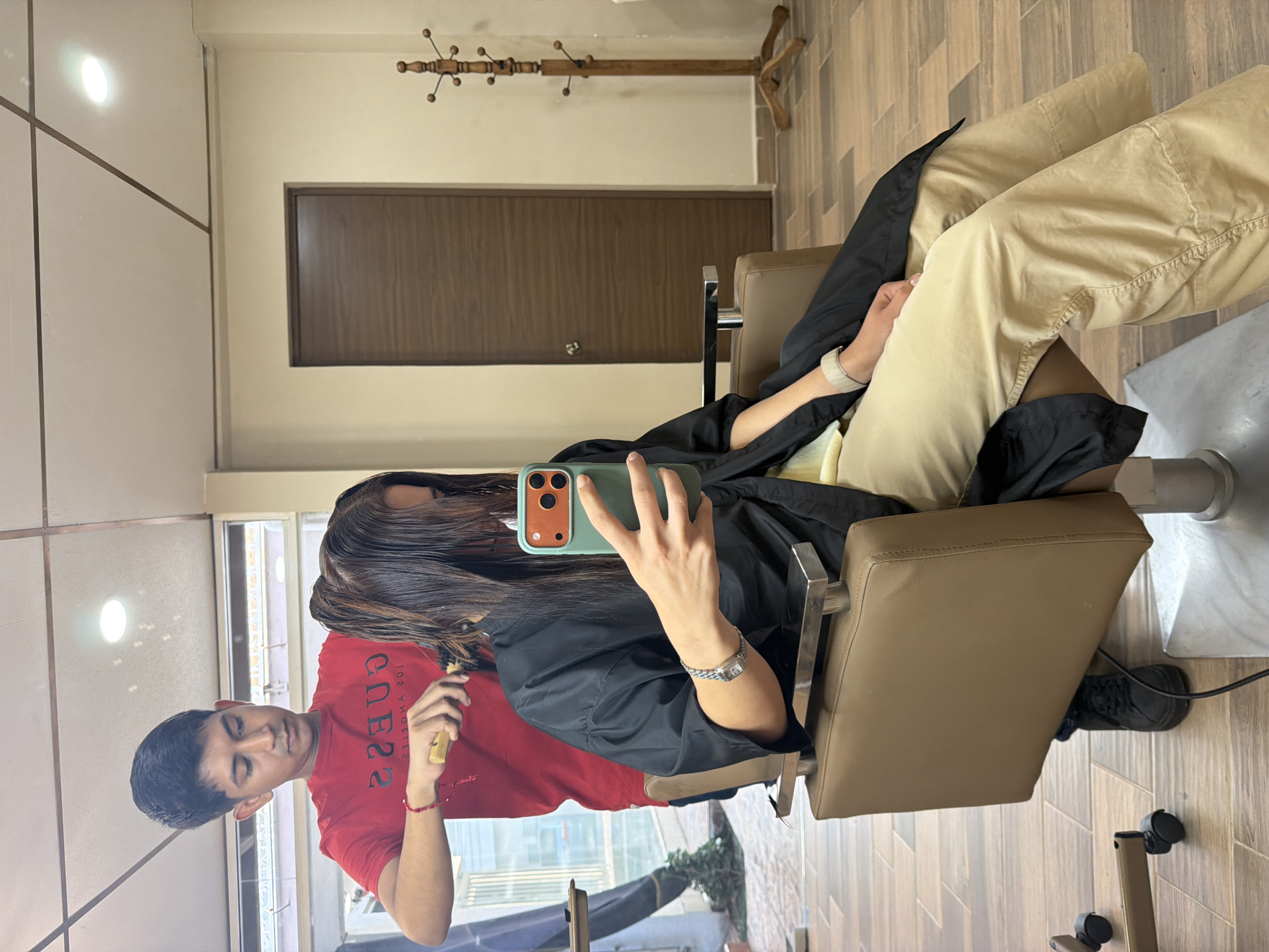 A woman wearing beige pants and a black salon cape is seated in a tan salon chair, holding a green phone, taking a photo or video. The location appears to be a salon in Villahermosa, Mexico, as can be seen from the reflections in the floor-to-ceiling window. A barber, wearing a red "Guess Los Angeles" t-shirt, is standing next to the woman, using a comb to style her hair. The flooring in the salon is wood-patterned, and the walls are a light beige color. A dark-brown door is visible in the background along with a wooden coat rack. There are ceiling lights visible in the mirror.
