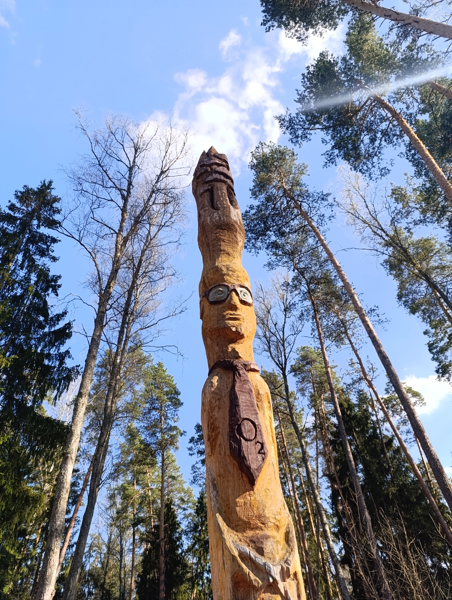 The image shows a tall, wooden statue in a forest in Unknown, Unknown. The statue is carved to resemble a person wearing glasses and a tie with the symbol "O2" on it. The statue is surrounded by tall trees, some with leaves and some bare. The sky is blue with a few clouds. The perspective is from a low angle, looking up at the statue and the surrounding trees.