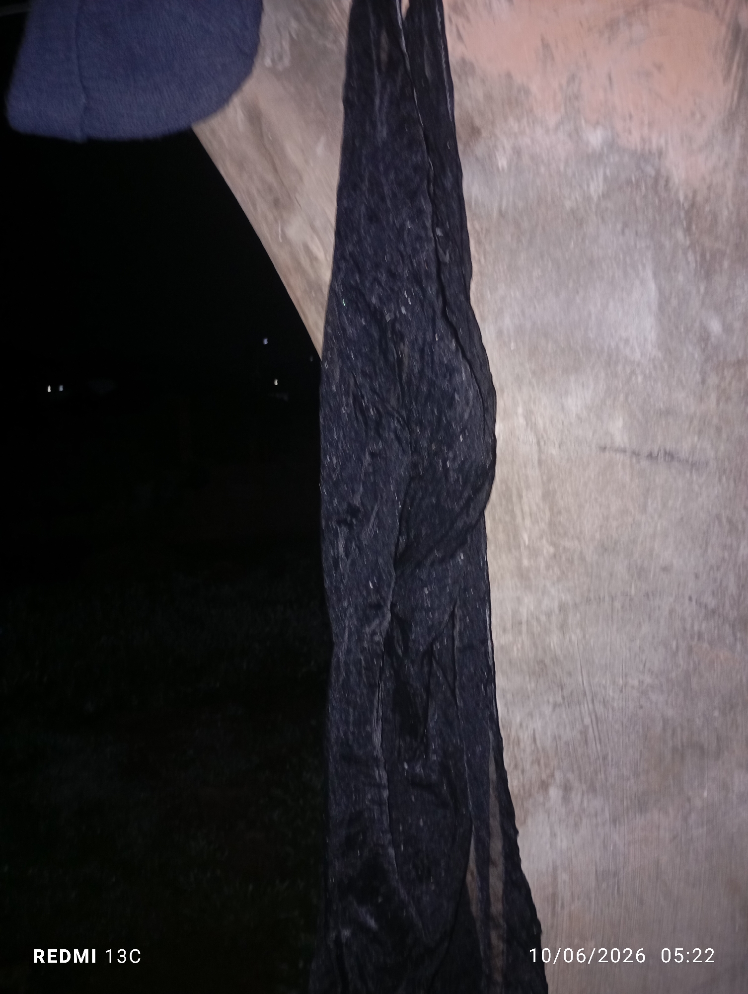A black, sheer fabric is draped over a wooden beam. To the left, a dark, blurred background suggests a nighttime setting with distant lights. A dark blue beanie is partially visible at the top left. The bottom right corner displays a timestamp "10/06/2026 05:22" and "REDMI 13C." The textured background is a light-colored, possibly concrete or rough plaster wall. This scene is not identifiable as being in Jos, Nigeria, or any specific urban location.