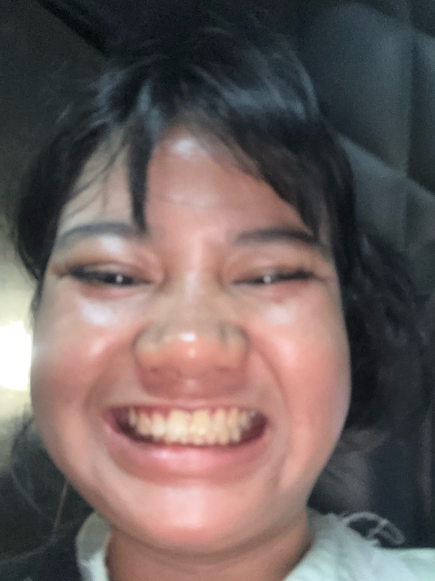 A blurry, close-up, over-the-shoulder selfie of a young woman with dark hair and bangs is taken indoors, possibly in a car. The woman is smiling broadly, with her teeth visible. Her cheeks are puffed out, and her eyes are narrowed in a playful expression. The lighting is dim, with a bright light source on the left side of the frame, casting a glare on her skin. The background is dark and indistinct, suggesting an indoor or nighttime setting. No specific location in North Jakarta, Indonesia, can be determined from the image.