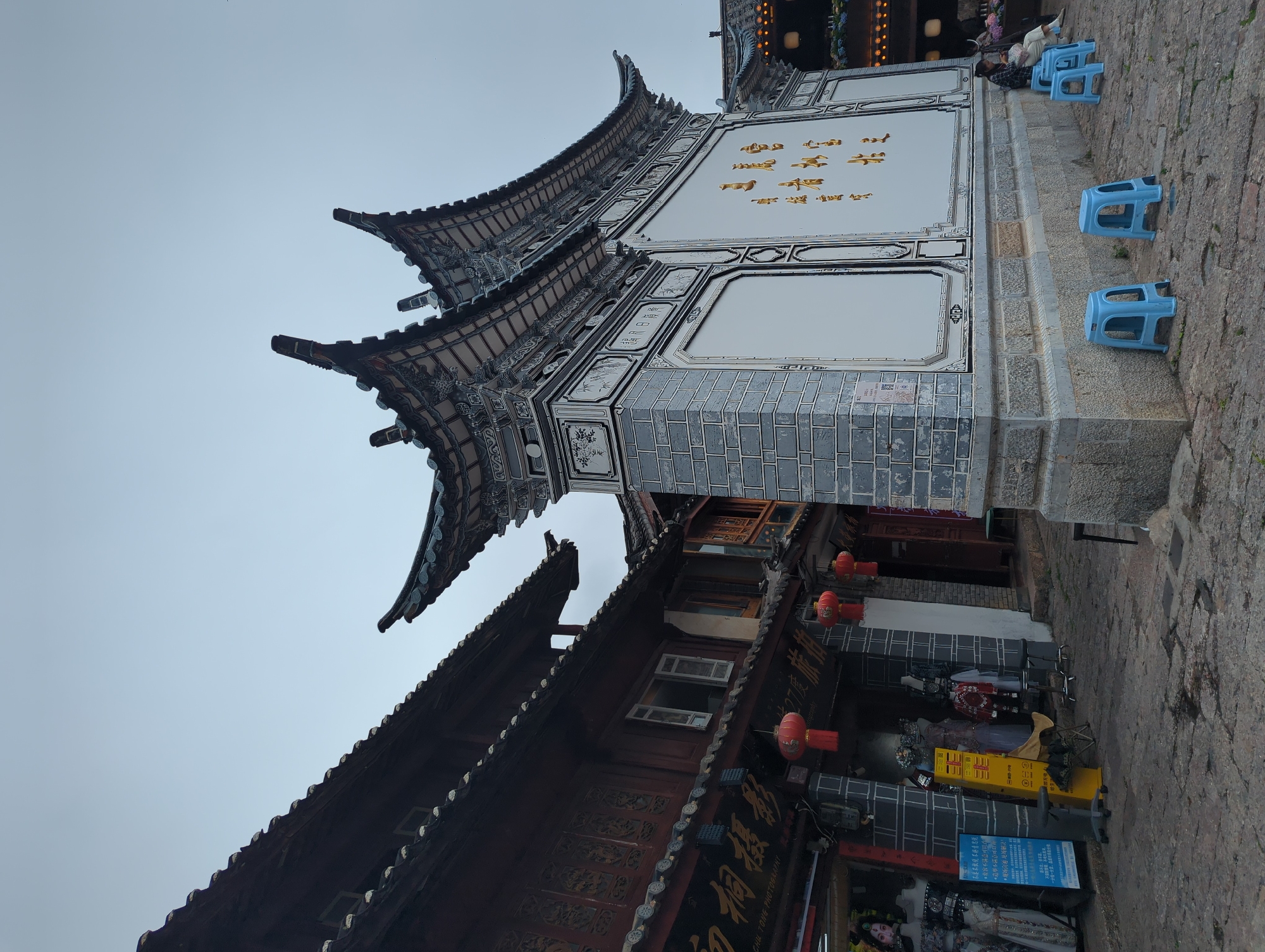 This image captures a street scene in Gucheng District, China, with a focus on traditional Chinese architecture. The foreground is dominated by the ornate, tiered roofs of buildings, characterized by their dark tiles and intricate detailing. The main building features a white facade adorned with golden Chinese characters and decorative panels, suggesting it might be a shop or a place of cultural significance.
Several bright blue plastic stools are arranged on a stone pathway in front of the building, some occupied by individuals. One person is seen sitting on a stool, appearing relaxed. The presence of these stools and the subdued lighting suggest it might be late afternoon or early evening. The sky is overcast, contributing to a muted atmosphere.
Below the main building, another structure with darker, wooden elements and a sloping roof is visible. Red lanterns hang from the buildings, adding pops of color to the scene. The cobblestone ground and the textured stone walls of the building reinforce the sense of a historic setting.
Visible text includes "CHU TONG PHOTOGRAPHY" and several lines of Chinese characters on signs and banners, some of which appear to be promotional or informational. The overall impression is of a quaint and culturally rich street in a Chinese town, likely a tourist area given the photography studio sign and the arrangement of seating.