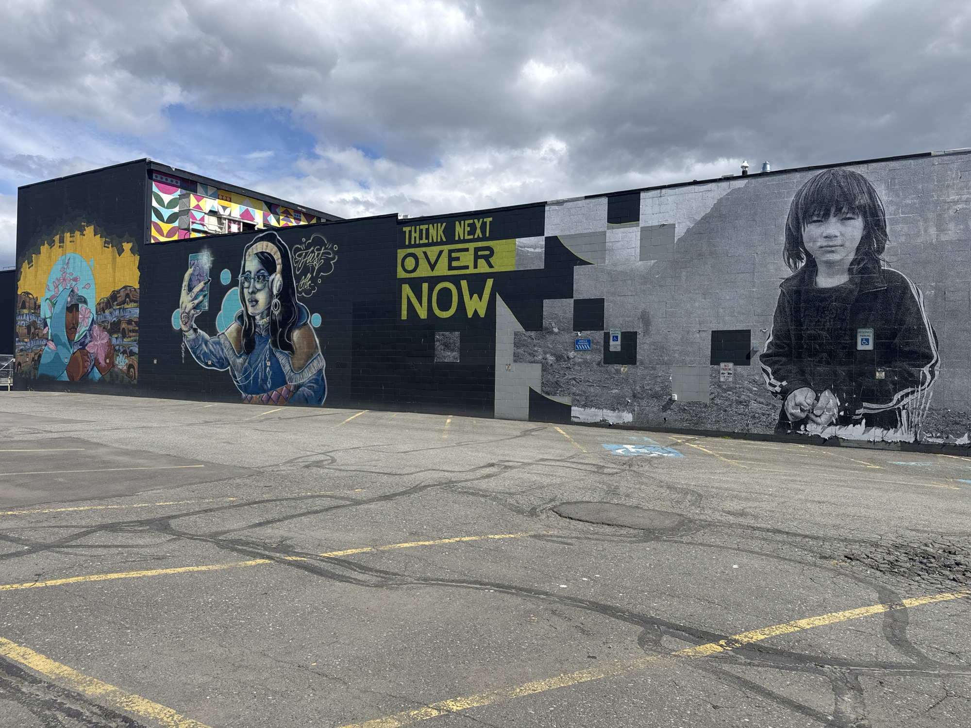 The image shows a mural on the side of a building in Anchorage, Alaska, United States. The mural depicts a variety of images, including portraits of people, abstract shapes, and text. The text reads, "Think Next Over Now." The parking lot in front of the building is cracked and has yellow lines. The sky is overcast.
