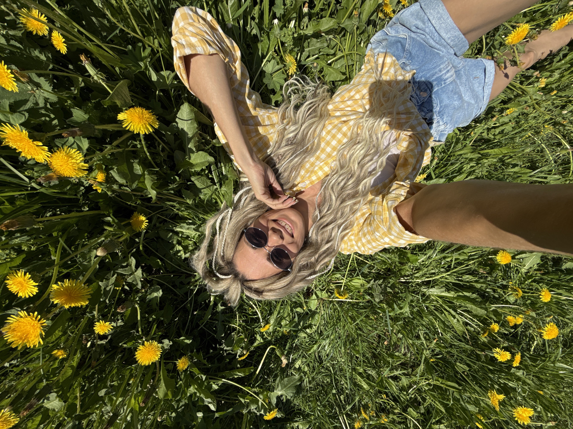 A woman is lying in a field of dandelions, taking a selfie. She is wearing a yellow and white checkered shirt and blue jean shorts. She has blonde, wavy hair with silver hair ties. She is wearing sunglasses. The background is a field of green grass and yellow dandelions. The location appears to be Rusanovo, Russia.