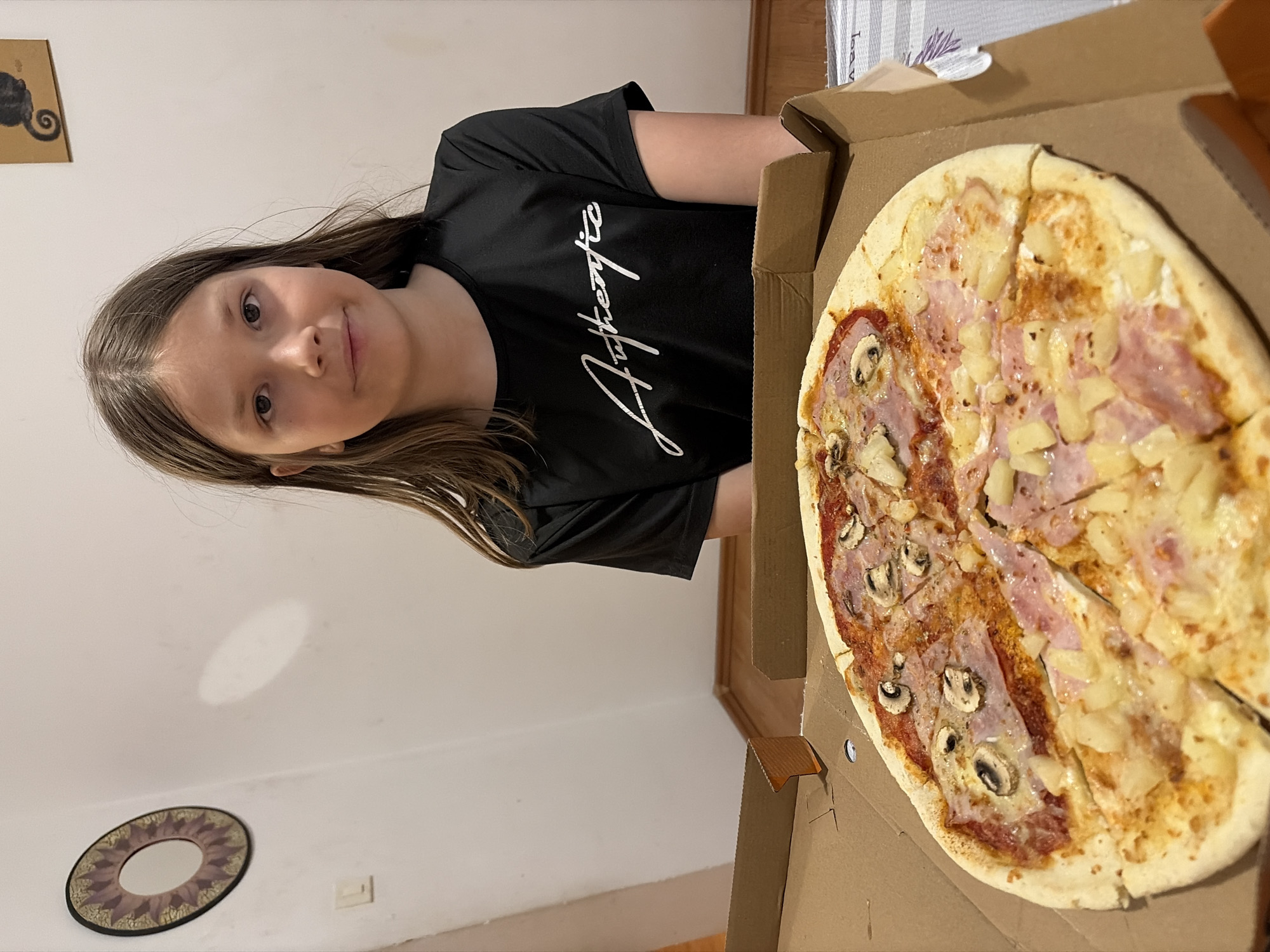 A young girl is pictured next to a pizza in a cardboard box. The pizza is half topped with mushrooms and ham, and the other half with ham and pineapple. The girl is wearing a black t-shirt with white writing. The setting appears to be indoors, possibly a home. There's a decorative item on the wall behind the girl and pizza. The photo does not provide any information to conclude that the location is in Budva, Montenegro.