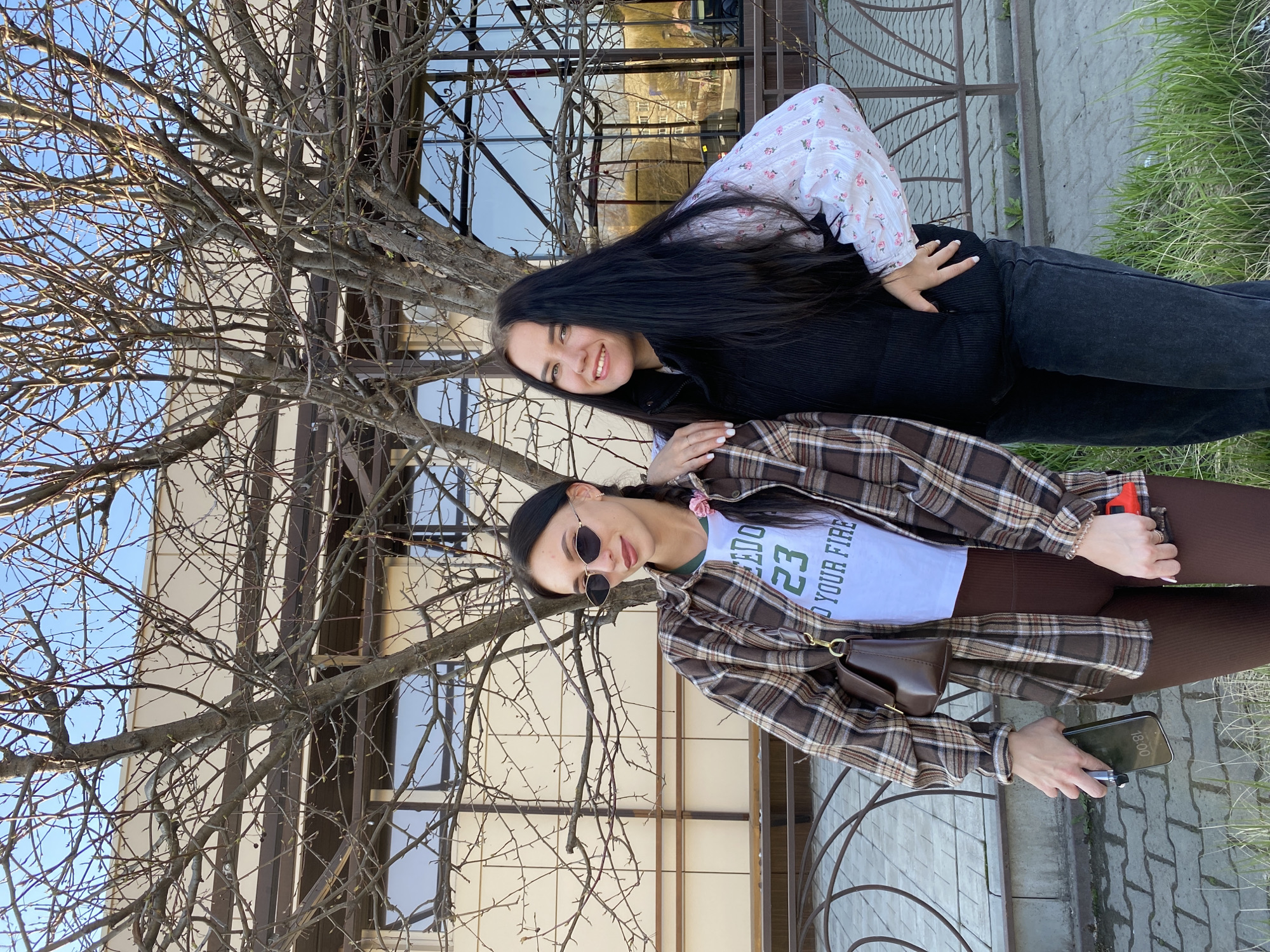 Two young women are posing for a photo outdoors. The background includes a building with large windows and a tree with bare branches. The woman on the top is smiling and has long dark hair. She wears a white blouse with small floral patterns and a black sweater. The woman on the bottom is wearing sunglasses, a brown plaid shirt, and brown leggings. She is holding a phone. The location appears to be in a city; specifically, Chaikovsky, Russia. The time on the phone is 18:00.