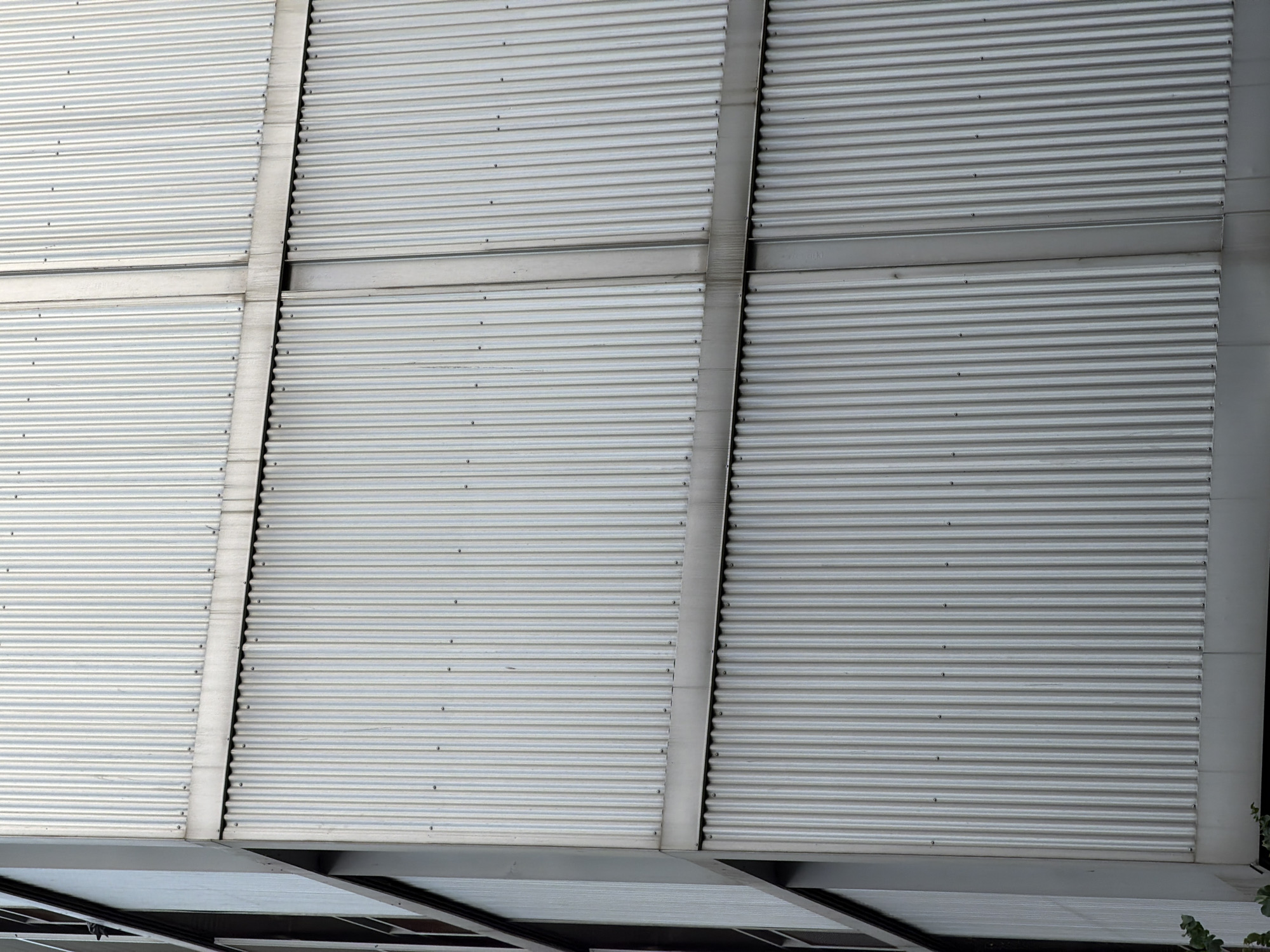 The image shows a close-up, eye-level view of a building's corrugated metal facade, likely in Barcelona, Spain. The facade is composed of vertical, ribbed metal panels in a silver-grey color. These panels are divided into sections by horizontal and vertical structural beams, also metallic. The texture of the metal is characterized by its repetitive parallel ridges, which create shadows and highlights, giving the surface a slightly textured and industrial appearance. There are no people visible in the frame. The lighting suggests it is daytime, with diffused light creating a generally even illumination across the surface, though some areas show subtle differences in brightness due to the corrugation and structural elements. The building's corner is visible on the left side of the image, revealing darker recessed sections which could be window frames or structural supports. A hint of greenery is visible at the very bottom left corner. The overall impression is one of modern, utilitarian architecture.