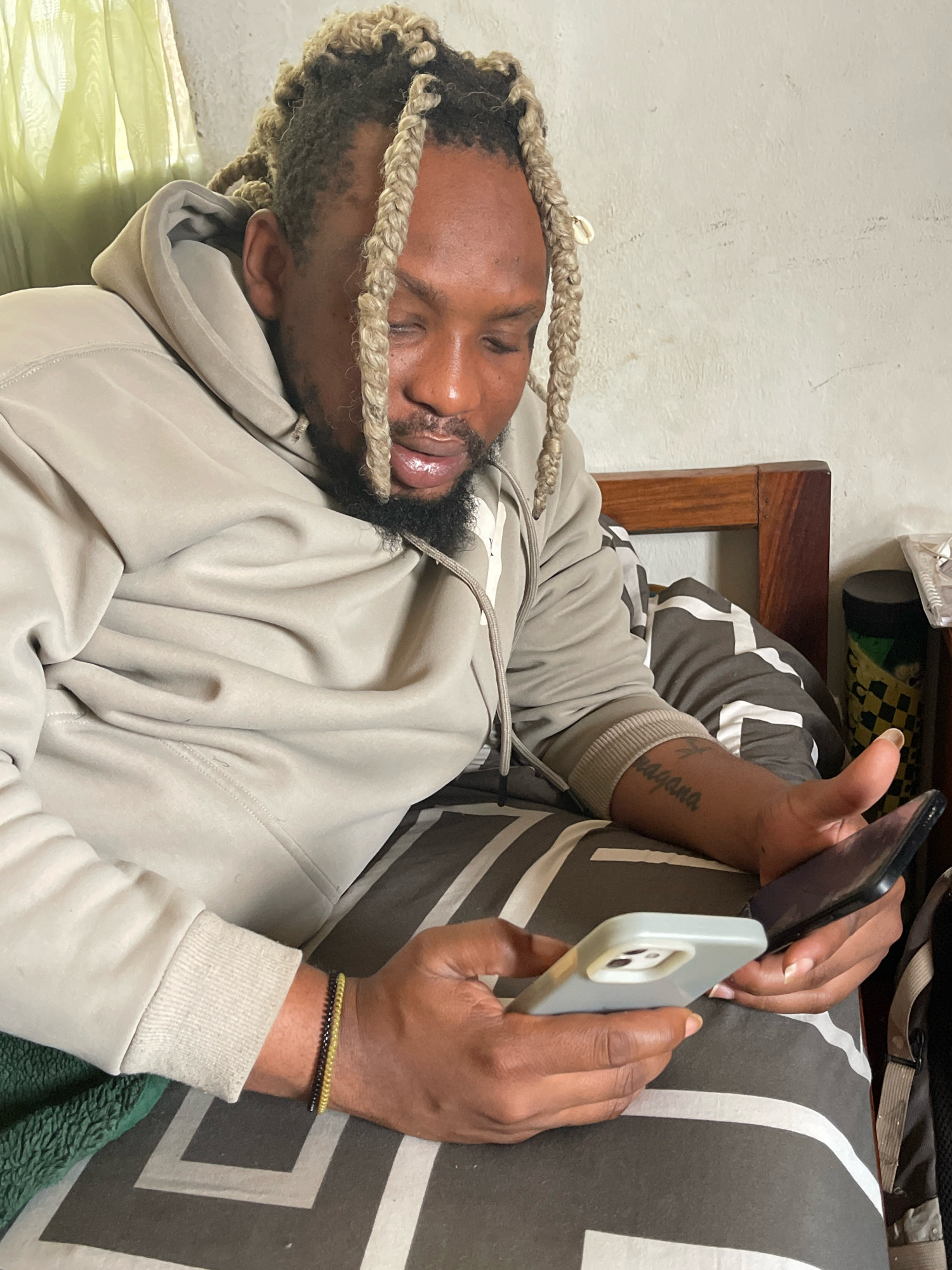 A medium shot shows a Black male with blonde braided hair, a beard, and a light gray hooded sweatshirt. He is lying down on a bed with a patterned duvet and is looking at two smartphones in his hands. His right arm, visible on his forearm, has a tattoo of the word "Vagava" and another word above it. He is wearing two beaded bracelets on his left wrist, one black and one yellow. The background shows a white wall, a wooden headboard, and a green curtain on the left. A dark cylinder and a plastic container are visible on a nightstand on the right. The scene is indoors, likely in a bedroom, in Jos, Nigeria.