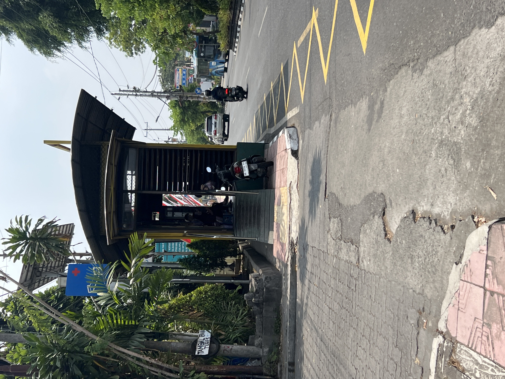 The image captures a street scene in Yogyakarta, Indonesia, specifically near a bus stop. In the foreground, a textured paved sidewalk transitions to asphalt road with yellow zigzag markings. A damaged section of the road is visible. To the left, a motorcycle is parked near the bus stop entrance, which features a yellow structure with a sloped, dark roof. A sign with "TAMBAL BAN TUBES" is mounted on a pole.
The bus stop itself has stairs leading up to an enclosed waiting area. Inside, two individuals are seated. Behind the bus stop, lush green trees and foliage are abundant. In the background, traffic moves along the road, including a white SUV and another motorcycle. A blue bus is also visible further down the street, alongside power lines and utility poles. A blue sign with a red cross symbol is mounted on a pole near the bus stop. The sky is overcast.