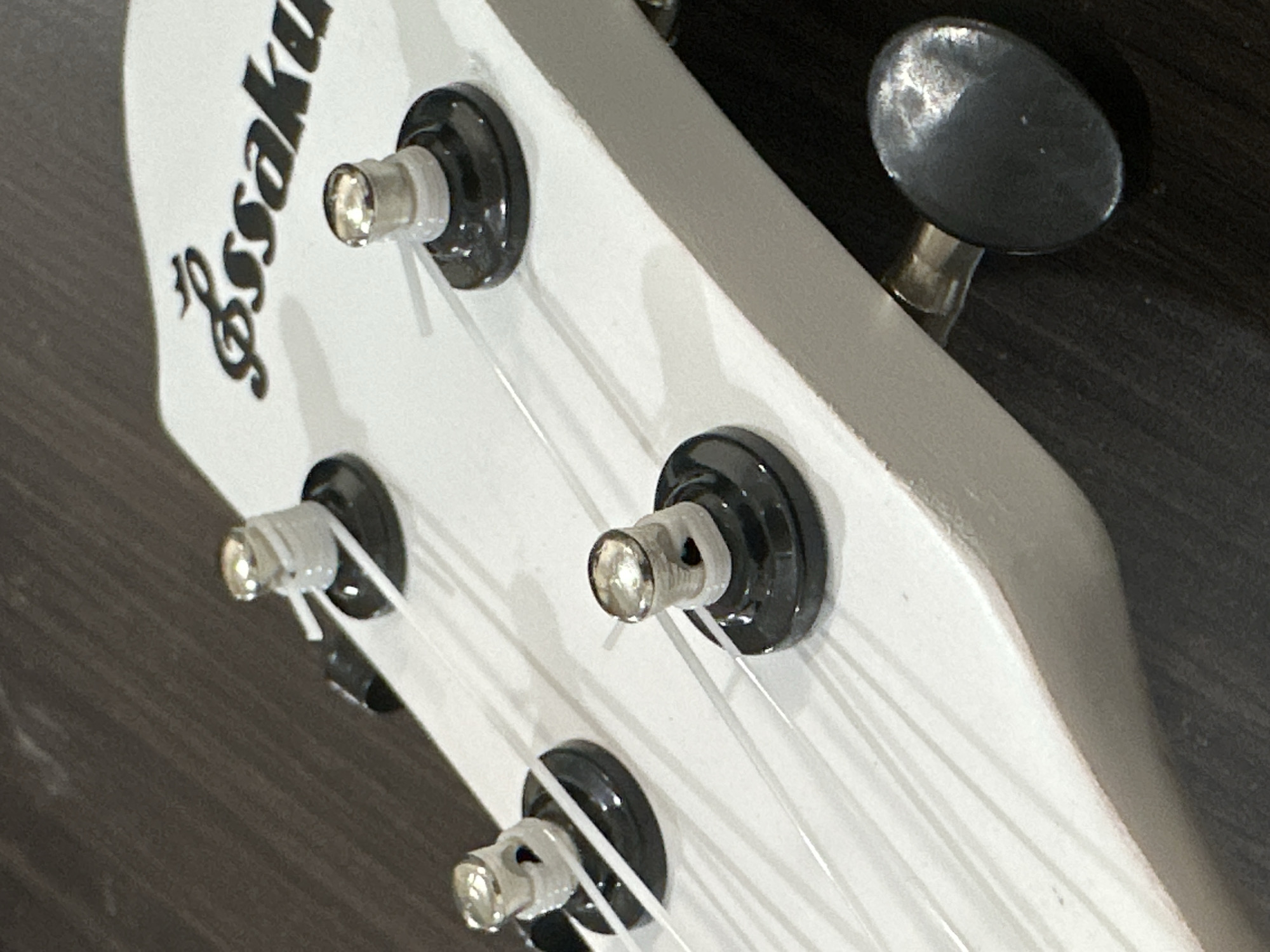 The image shows a close-up, high-angle shot of the headstock of a white ukulele. Four tuning pegs with black bases and silver caps are visible, along with their associated strings. The strings are thin and white, stretching across the frame. A black tuning key is also present on the right side of the image. The ukulele headstock features a black logo with cursive lettering that reads "Essakula". The background is a dark, textured surface, likely wood. There are no people or animals present in the image. The scene is indoors and well-lit, with the focus on the details of the ukulele's tuning mechanism and branding. There is no information or context that would allow for the determination of a specific location in Bulakrejo, Indonesia.