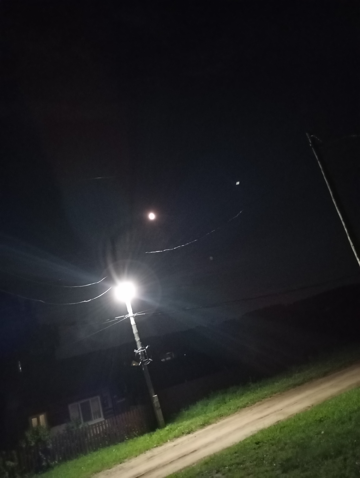 The image depicts a nighttime scene in the city of Foky, Russia, characterized by a dirt road with grass on either side. A street light illuminates the area, casting a bright light upwards and outwards. Above the street light, a faint moon is visible in the dark sky, along with some power lines. In the background, a wooden fence surrounds a property with a house that has illuminated windows. The overall impression is of a quiet, rural setting at night, with the artificial light of the street lamp and the natural light of the moon creating a stark contrast against the darkness.