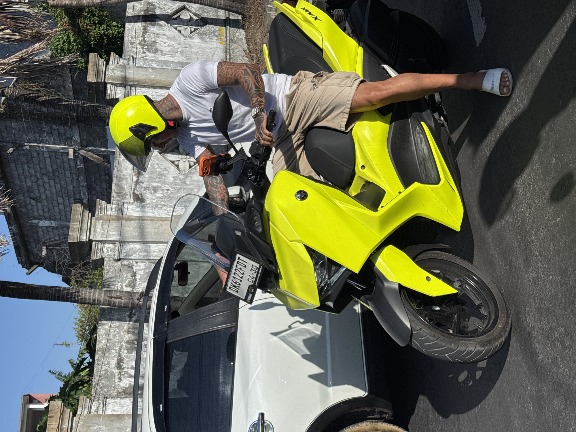 A man wearing a bright yellow helmet and a white t-shirt sits on a bright yellow scooter, parked next to a white car. He has tattoos on his arms and neck. The scooter has a license plate that reads "DK6122FDT 04.30". The scene appears to be in a city, possibly Denpasar, Indonesia, based on the style of architecture and vegetation, including palm trees in the background. The ground is paved and there are shadows cast by the vehicles and a nearby building. The weather is sunny.