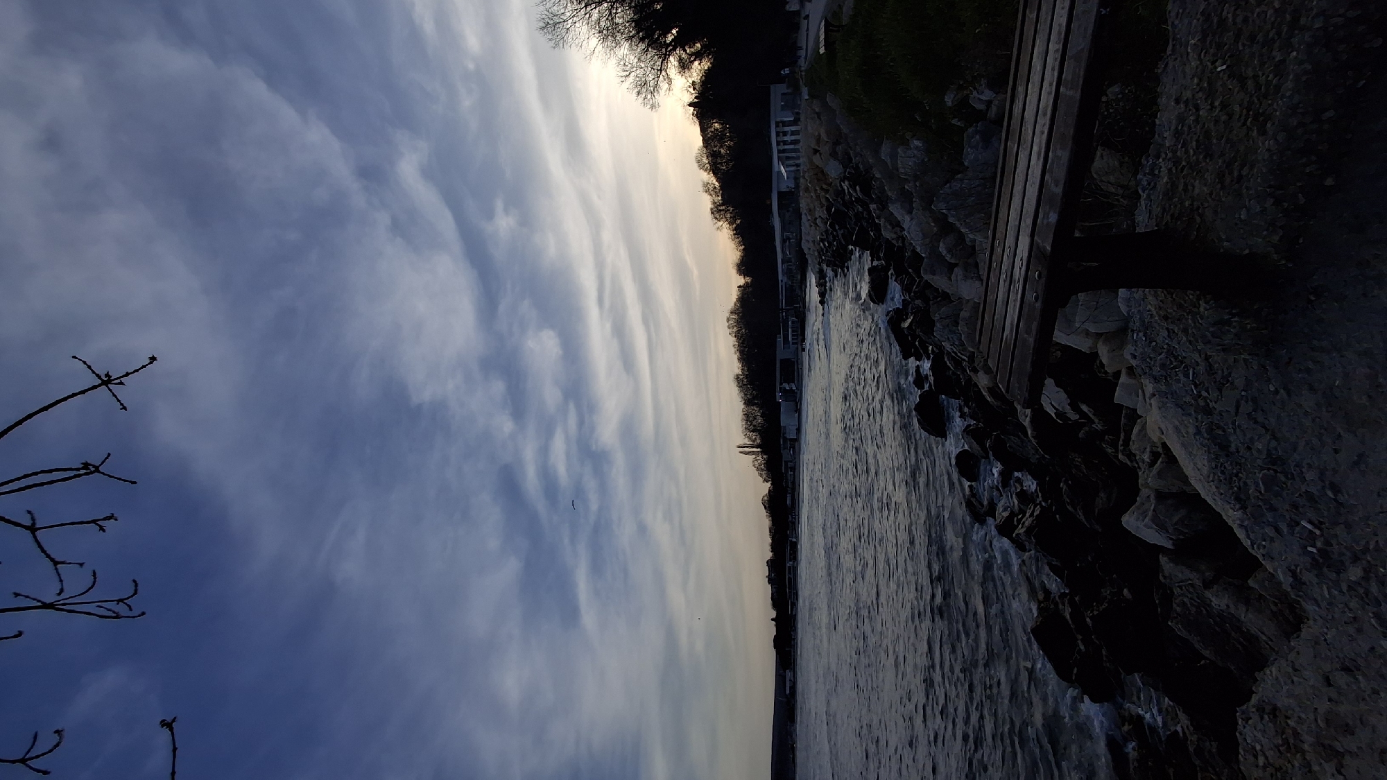 The image depicts a coastal scene in Varna, Bulgaria, at either sunrise or sunset. The sky is predominantly blue with white and grey clouds. A thin strip of light from the setting sun is visible along the horizon. The shoreline consists of a rocky embankment, where a wooden bench is partially visible on the right side of the frame. The dark water of the sea or bay is situated between the sky and shoreline. Bare branches of a tree extend from the lower left corner into the sky. Buildings are dimly visible along the coast.