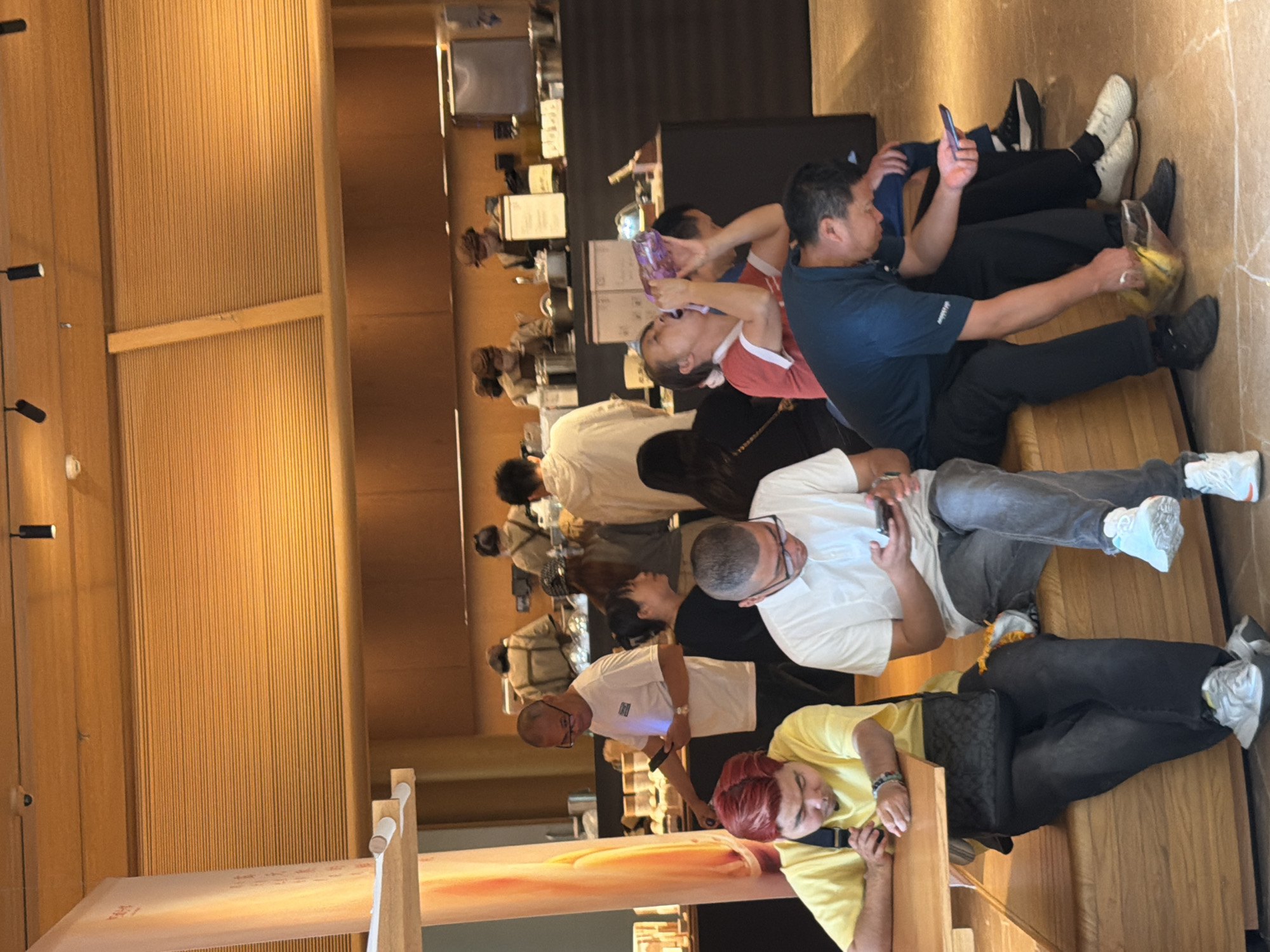 In Yuzhong District, China, a group of people are gathered in what appears to be a cafe or food establishment with a warm, wooden interior. In the foreground, a man with red hair, wearing a yellow t-shirt and dark pants, is seated at a wooden table, looking down at his phone. To his right, a man in a white polo shirt and gray jeans is also looking at his phone. Further to the right, a man in a dark blue shirt and black pants is holding a yellow bag or container. In the background, several other individuals are visible, some interacting with staff behind a counter. The overall atmosphere suggests a casual gathering or a brief stop for refreshments.