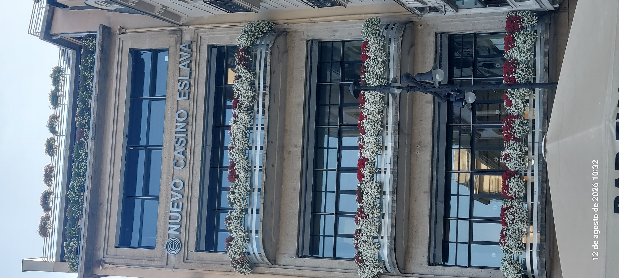 The image shows the facade of the Nuevo Casino Eslava building in Pamplona, Spain. The building has three floors visible, each with large, dark-paned windows. The upper floors feature balconies adorned with rows of red and white flowers. A street lamp with multiple bulbs stands in the foreground to the right of the building. The sky above is clear and blue. Text overlays at the bottom of the image indicate a date and time: "12 de agosto de 2026 10:32".