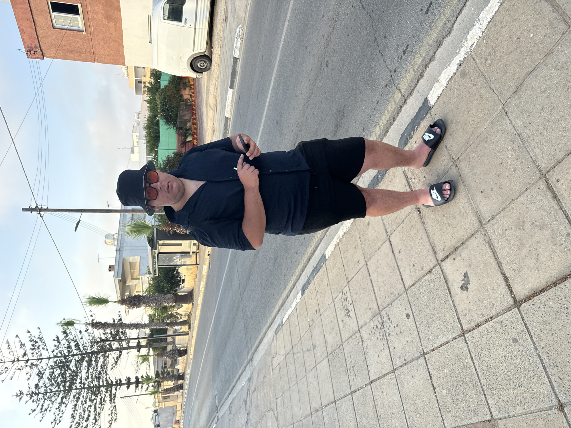 A man stands on a sidewalk next to a street in Xylofagou, Cyprus. He is wearing a dark bucket hat, red sunglasses, a dark short-sleeved button-up shirt, and black shorts. He is holding a smartphone in his right hand and a cigarette in his left. The man is wearing black Nike slides on his feet. Behind him, a white van is parked on the side of the road, and several buildings and palm trees are visible. The sky is clear with a few clouds. The street appears to be a two-lane road with a sidewalk.