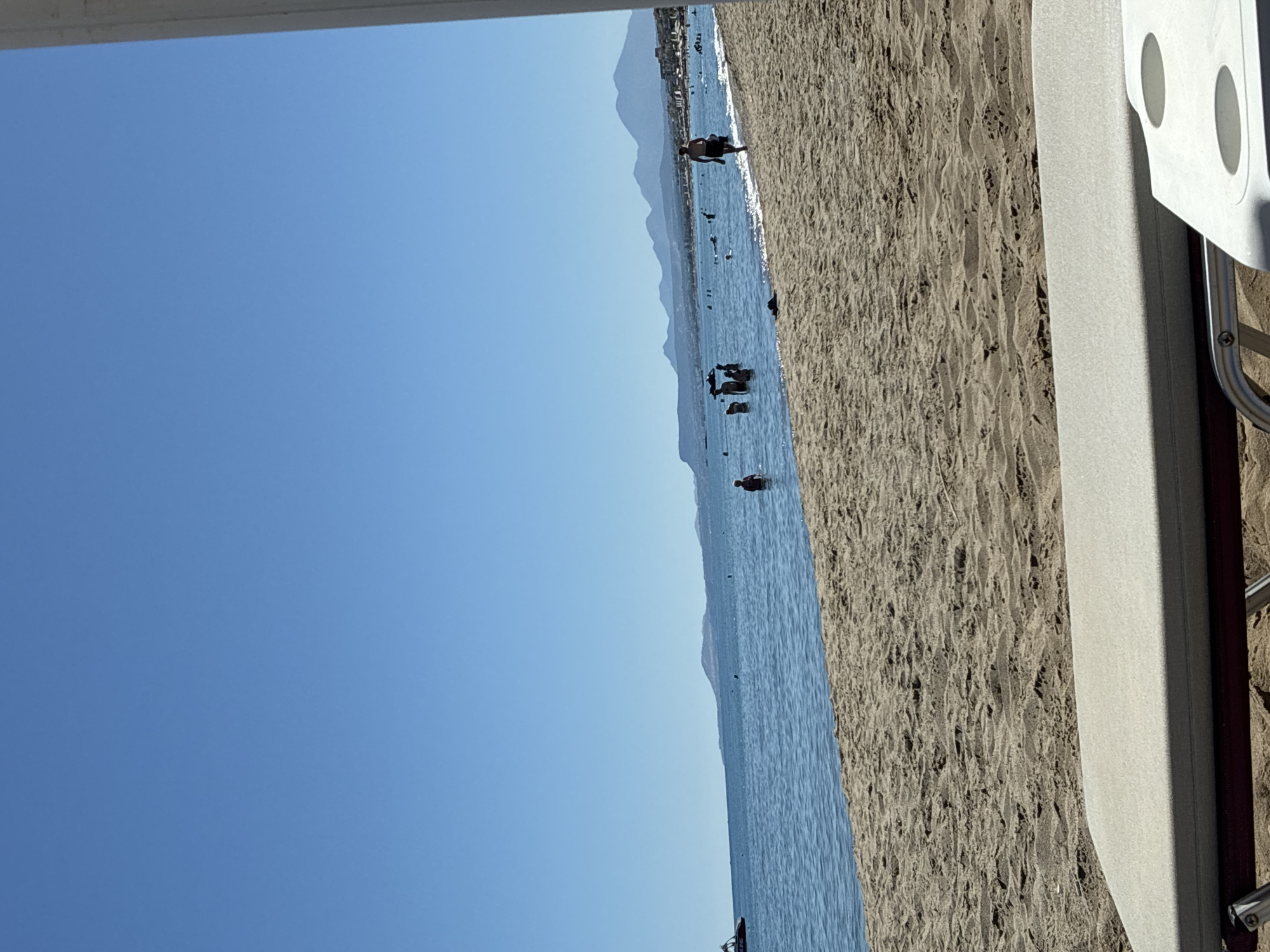 The image depicts a sunny day at a beach in Rethymno, Greece. In the foreground, a partially visible beach lounger is set on the sand. The midground features the sandy beach leading to the calm, blue sea, where several people are swimming and wading. A jet ski is visible in the far left of the water. In the background, a picturesque coastline with buildings and mountains stretches across the horizon under a clear blue sky.