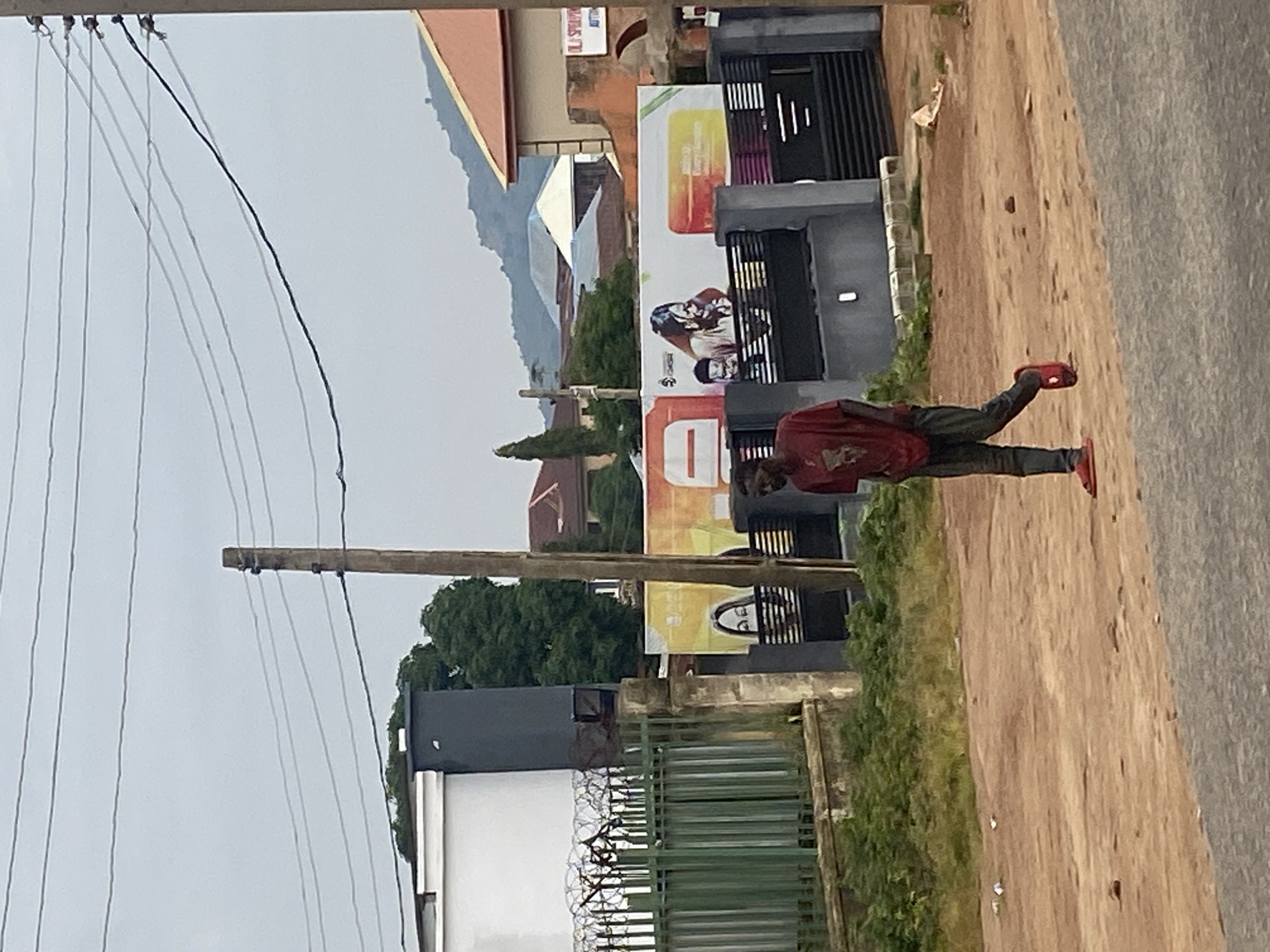 An outdoor, eye-level, medium shot captures a person walking on a dirt road in front of a fenced property with a billboard. The individual, a dark-skinned male, wears a red t-shirt and dark pants. Red sandals are on his feet. He is in mid-stride, facing left, with his head down. The dirt road is textured with small pebbles and debris. To the left, a green metal fence with barbed wire atop separates the road from a property with a dark gray, rectangular structure and a white section. Behind the fence, dense green foliage is visible. A large billboard featuring images of two people, one with long dark hair, and text, is mounted behind a dark metal gate, on a gray wall. Power lines crisscross the sky above. In the background, various buildings with orange and brown roofs are scattered amongst green trees, with hills in the far distance. The sky is overcast with a grayish hue. The scene appears to be in Jos, Nigeria, based on typical urban landscapes of the region.