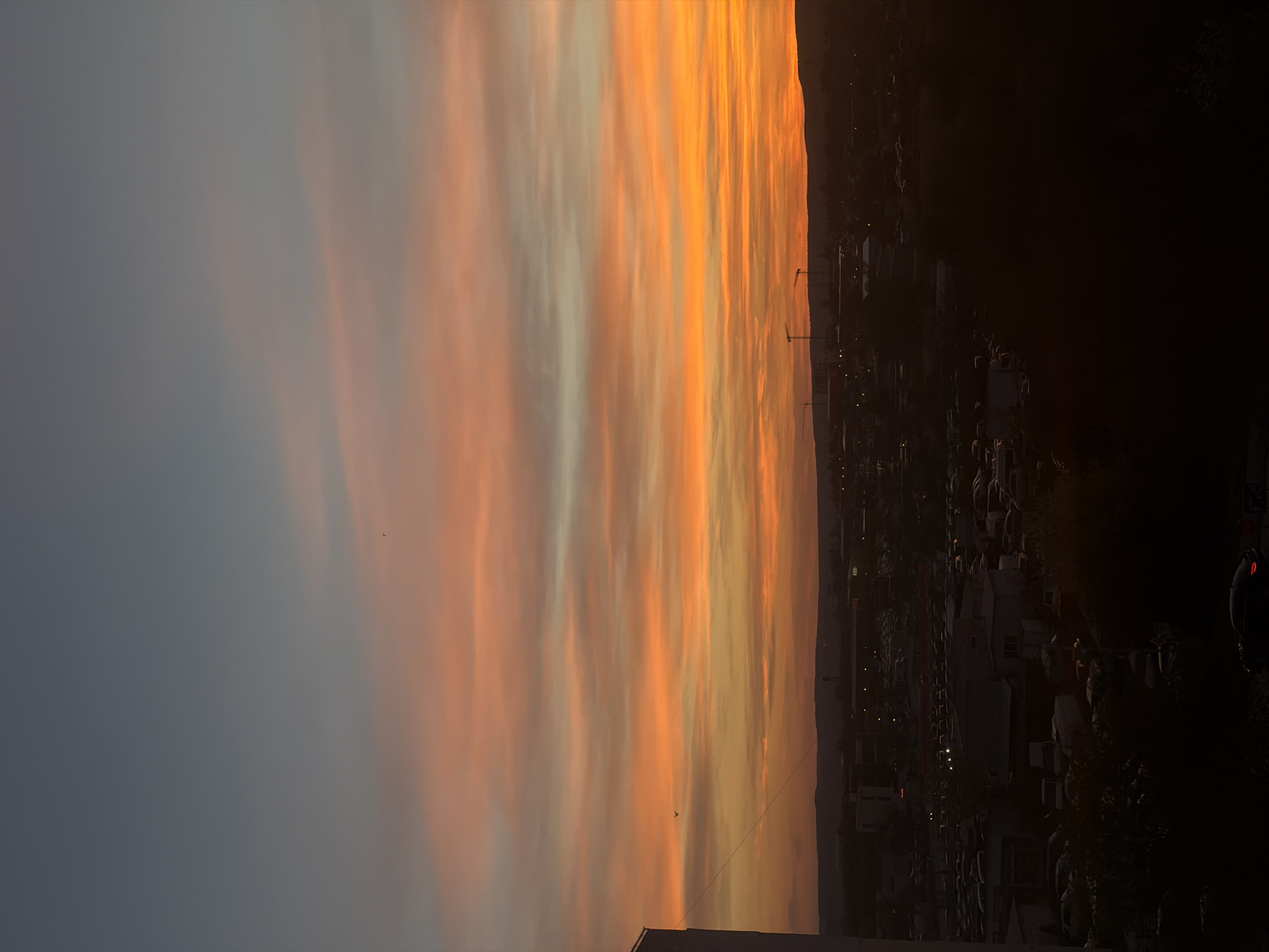 A panoramic view captures a vibrant sunset over a city, likely Varna, Bulgaria, seen from an elevated perspective. The sky dominates the upper two-thirds of the image, filled with dramatic, streaky clouds. Near the horizon, these clouds glow with intense shades of orange, yellow, and red, indicative of the setting sun. Further up and to the left, the cloud colors transition into softer peach, light gray, and finally a muted blue-gray in the upper left quadrant. The clouds run in elongated, horizontal patterns across the sky.
Below the colorful sky, a dark, indistinct silhouette of distant hills or mountains marks the horizon. The mid-ground and foreground present an urban landscape cast mostly in deep shadow due to the strong backlighting from the sunset. Numerous buildings appear as dark shapes, some with faint internal or street lights visible as small, bright points. Several tall construction cranes are clearly silhouetted against the brightly lit portion of the sky, suggesting ongoing development. Dark masses of vegetation and indistinct shapes of vehicles can also be seen within the shadowed cityscape.
The image portrays the serene yet striking beauty of dusk, characterized by the stark contrast between the warm, illuminated sky and the cool, shadowed ground. No people are visible, and no text is present in the scene. The time of day is clearly evening, during sunset.