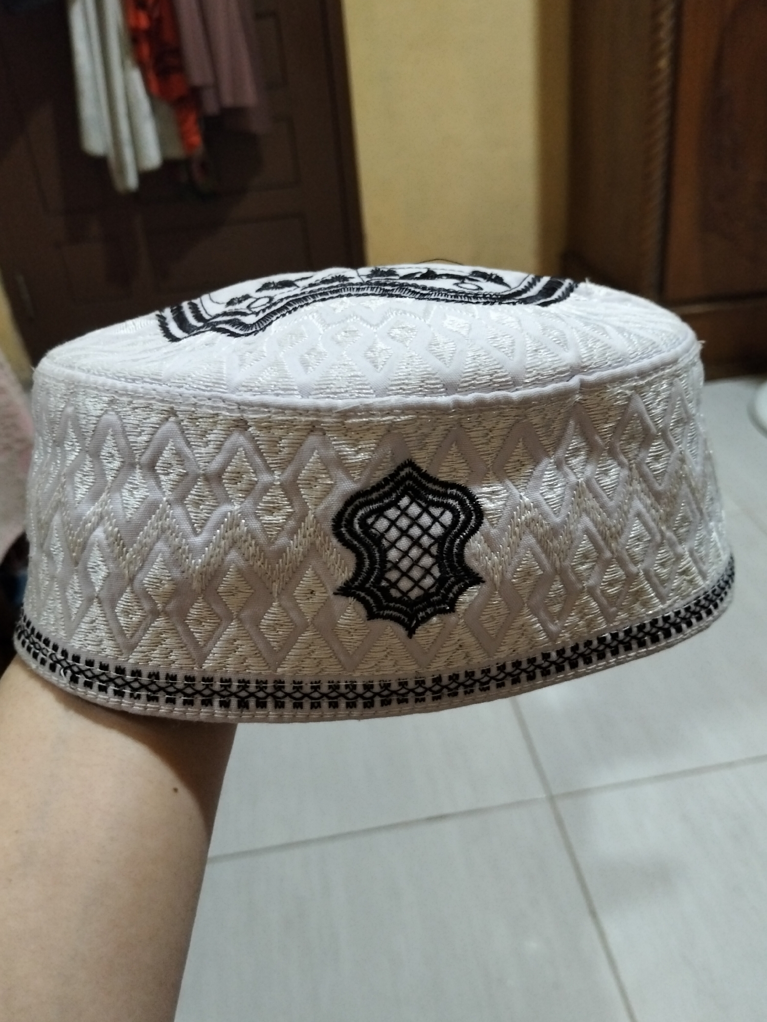 A white kufi cap with black embroidery is held up on a person's arm. The kufi has a diamond-shaped pattern and a decorative motif on the front. The background is blurred, showing a closet with hanging clothes and a wall. The scene appears to be indoors.