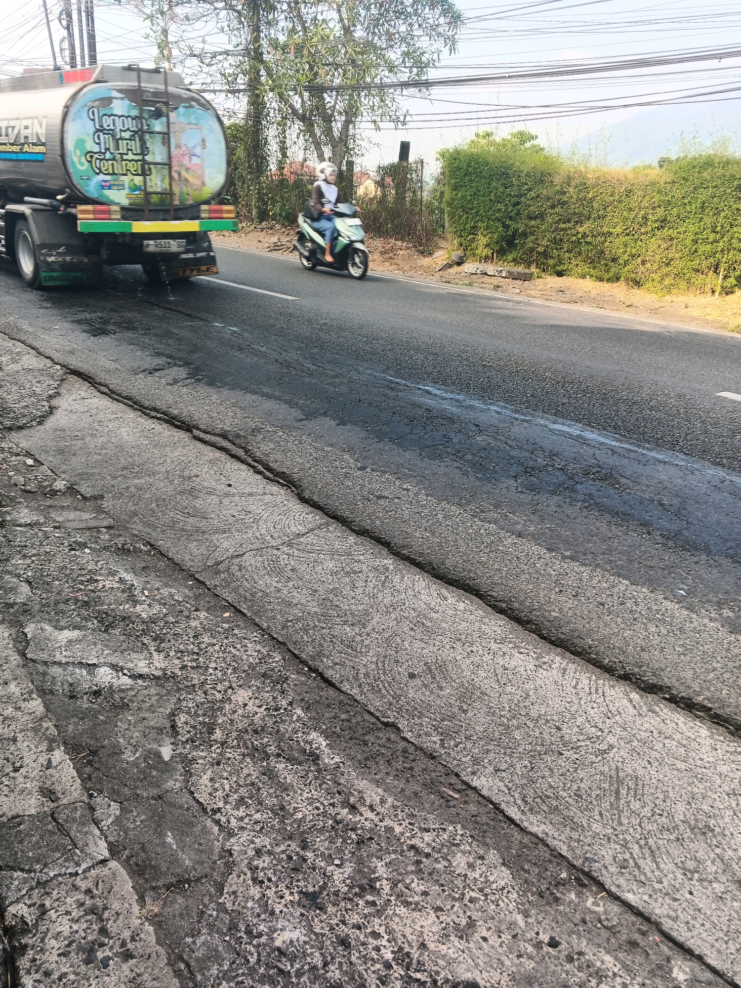 The image depicts a street scene in Ungaran, Indonesia, characterized by damaged road surfaces. In the foreground, the road shows significant cracking and deterioration, with patches of exposed aggregate and unevenness. To the right, the road is paved with asphalt, also exhibiting cracks and some wet, oily patches, suggesting recent activity or leakage.
Further down the road, a water tanker truck is visible on the left. The truck's tank has a colorful mural on its side, depicting what appears to be a cartoonish scene. The truck is in motion, moving away from the camera.
On the right side of the road, a person wearing a helmet rides a scooter. The rider is dressed in casual clothing. The scooter is also in motion, moving in the same direction as the truck.
The background features a dense green hedge and trees, with utility poles and wires overhead. The sky is bright and clear, indicating daylight. The overall scene captures a typical Indonesian road with the presence of vehicles and the common issue of road maintenance.