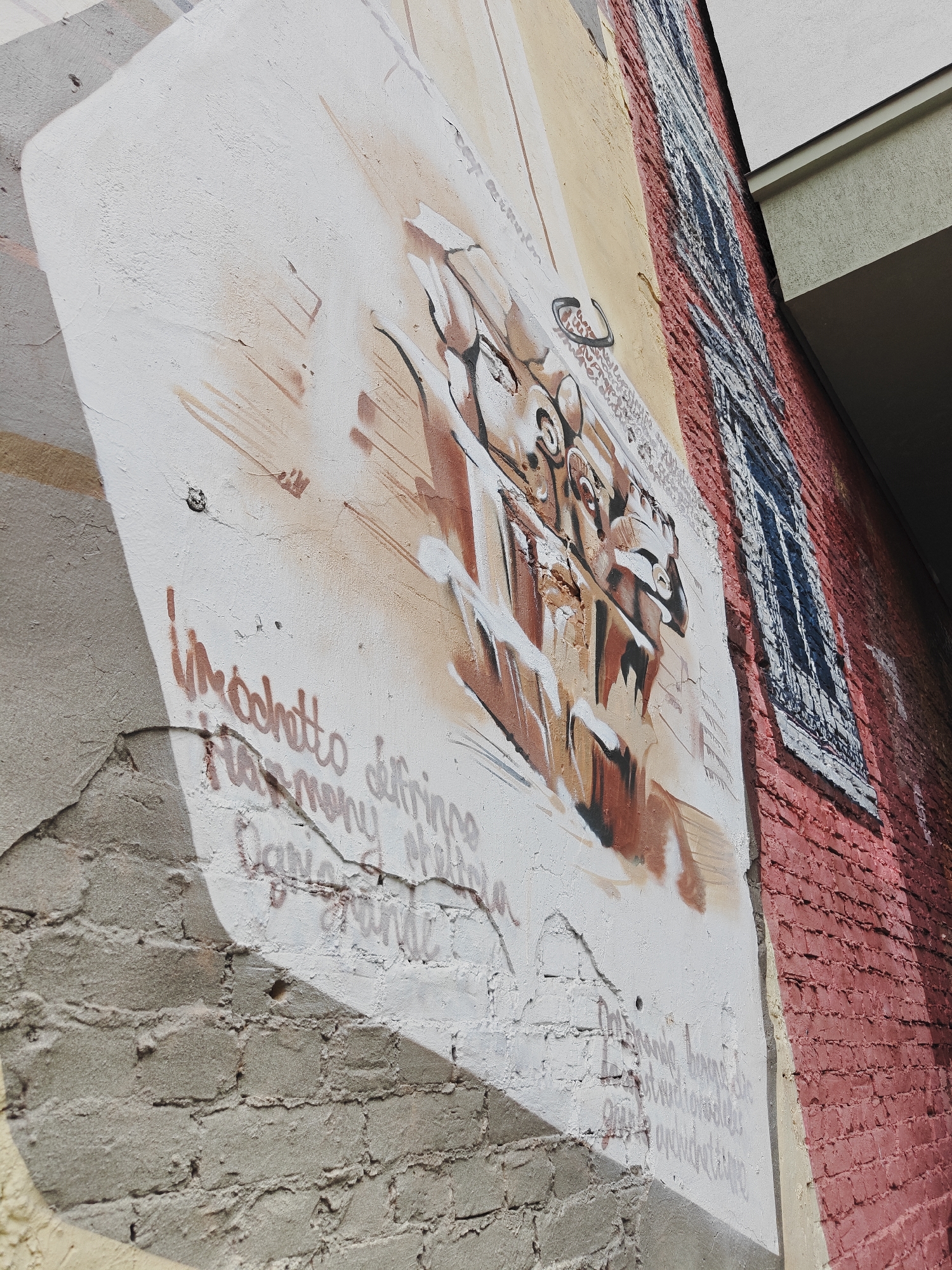 This is a medium-shot, eye-level, exterior, daytime photograph of a section of a building's facade decorated with graffiti art in Saint Petersburg, Russia. The wall is divided into different sections with varying textures and colors. The main focus is a large mural painted on a white, textured surface, featuring abstract figures and lettering in shades of brown, black, and white. To the right of the mural, a section of the wall is made of red brick, with a painted depiction of a window with a dark blue and white grate. The upper right portion of the image shows a light-colored overhang and part of another building. The graffiti art includes what appears to be stylized lettering in a reddish-brown color that reads, "Immohotto Harmony delfrinne" and other indecipherable script. Further down, lighter gray script is visible. The overall condition of the wall shows signs of age and wear, with cracks and chipped paint evident. The lighting suggests a sunny day.