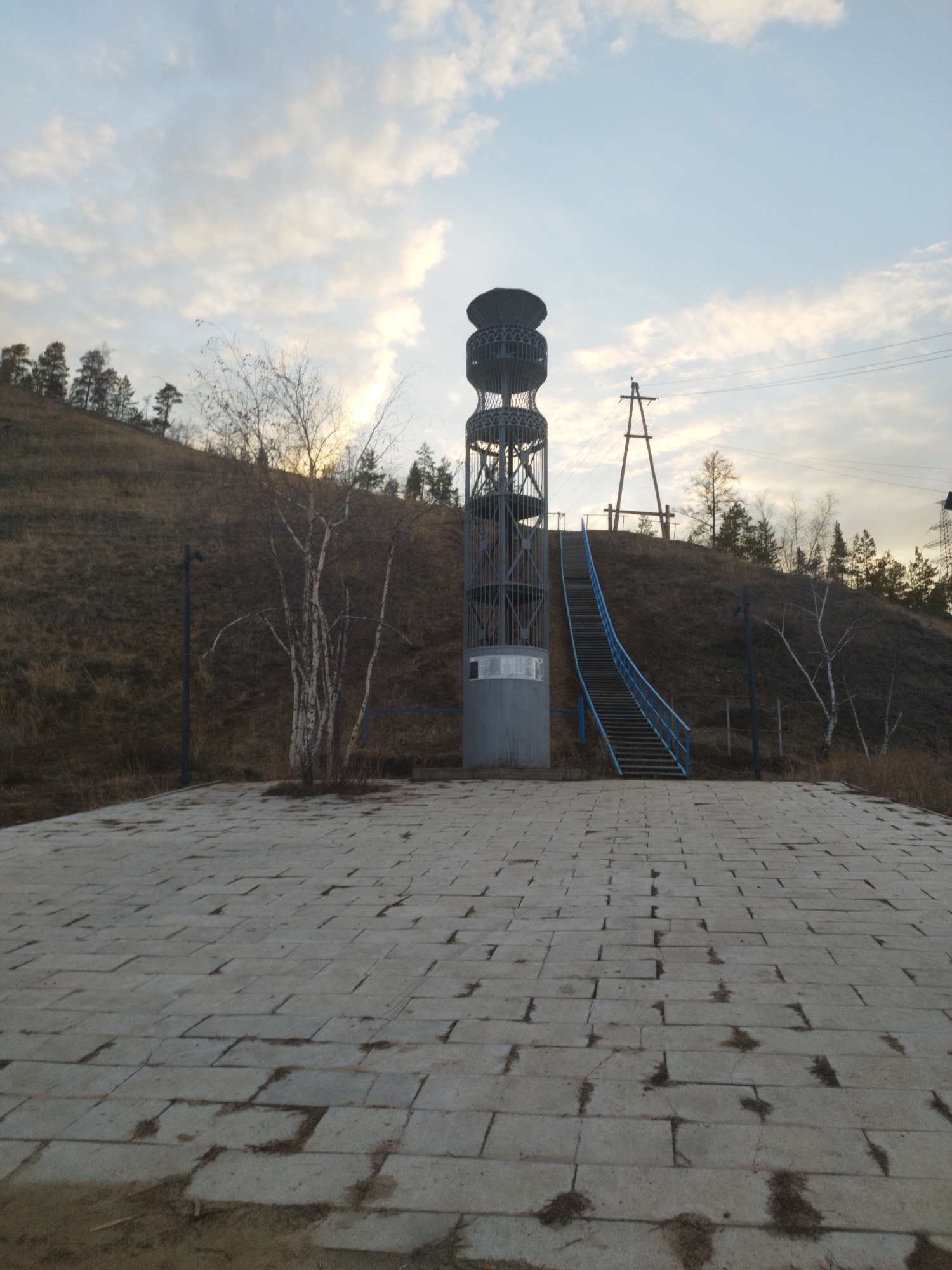 The image depicts a landscape featuring a metal observation tower with a spiral staircase, located on a hill in what appears to be an open area. The tower is constructed with a cylindrical base, supporting a multi-tiered open-cage structure capped with a disc-shaped top. A set of stairs with blue railings leads up the hillside behind the tower. In the background, an electrical power line is strung across the hillside supported by poles. The ground in the foreground is paved with rectangular stone blocks. Sparse vegetation is seen on the hillside, including trees with bare branches suggesting it may be late autumn or early spring. The sky is overcast with a pale, diffused light. The image is of the Mammoth Observation Deck and Tourist Complex in Yakutsk, Russia.