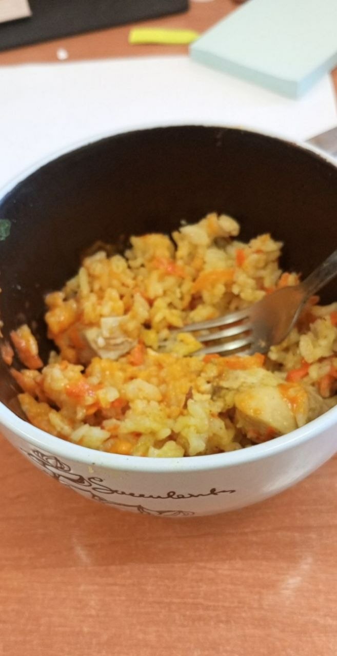 A close-up shot depicts a bowl of food, likely rice pilaf with chicken and carrots, being eaten with a fork. The bowl is white with a dark rim and decorative floral patterns. The food appears to be sitting on a wooden or wood-like table. In the background, slightly out of focus, there are items including a black object, a yellow marker and a small stack of papers. There is no evidence to specify Minsk or Belarus as the location.