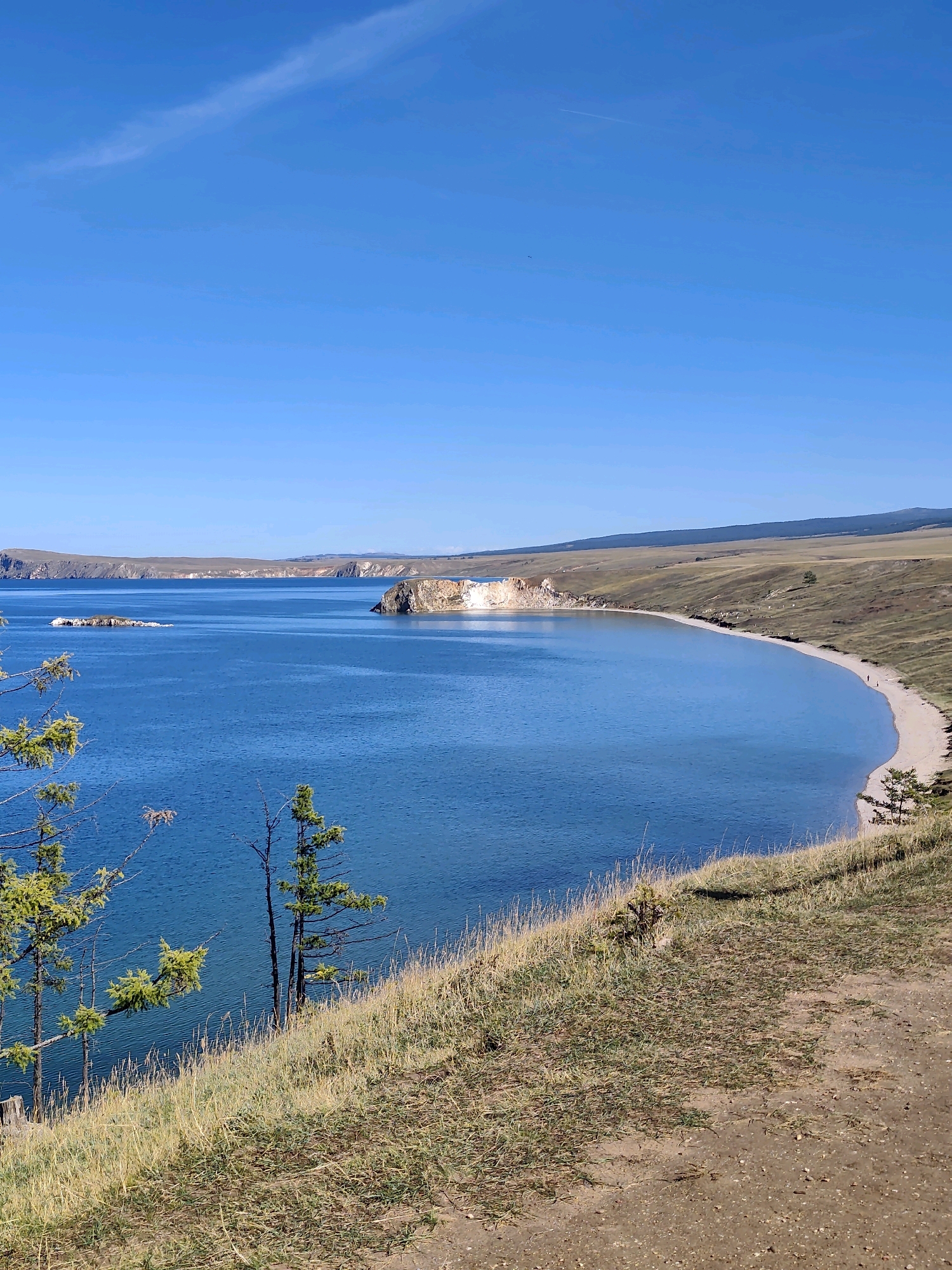 The image depicts a panoramic view of a coastline on a clear, sunny day. The foreground features dry, golden grass and dirt path, suggesting a natural, somewhat arid landscape. Several sparse evergreen trees are visible on the grassy slope, some appearing thin and wind-swept.
In the midground, a wide expanse of deep blue water stretches out, with gentle ripples indicating calm conditions. The water meets a sandy beach that curves along the shoreline. Further in the distance, the coastline is marked by low, rolling hills covered in similar dry vegetation. A prominent rocky outcrop, with a lighter, possibly white or cream-colored face, juts out from the land into the water on the right side of the bay. Further to the left, a smaller, rocky island is visible in the water.
The sky is a vibrant, cloudless blue, with only a faint, wispy contrail high in the upper left quadrant. The overall scene conveys a sense of vastness and natural beauty. The mention of Kharantsy, Russia, suggests this is likely a remote or natural area within or near that location. There are a few small, distant figures visible on the beach, suggesting human presence but not significant activity. The image appears to be taken from an elevated vantage point, looking down and across the bay.