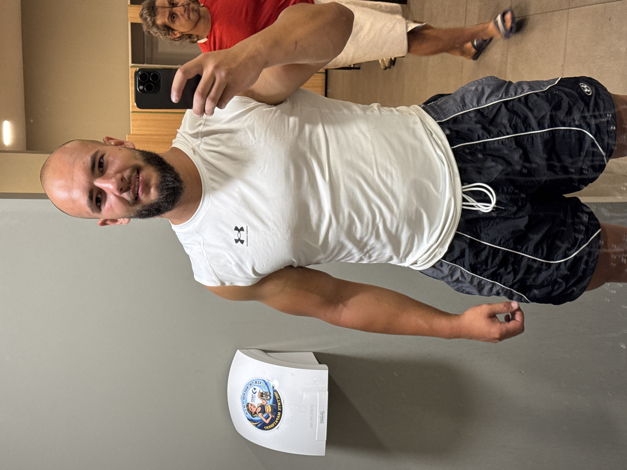 A bald man with a beard, wearing a white sleeveless Under Armour shirt and black shorts with white trim, takes a mirror selfie in a gym locker room. Another man in a red shirt and white towel is visible in the background. A white hand dryer with a circular sticker featuring a cartoon woman is mounted on the grey wall to the left. The floor is tiled. The image appears to be taken in the city of Sofia, Bulgaria.