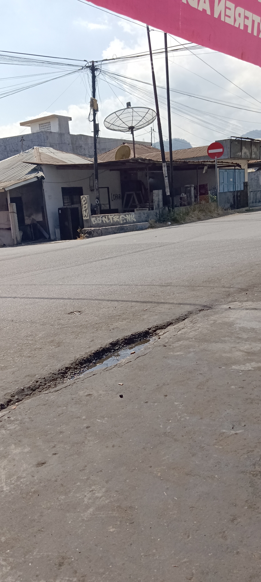 The image depicts a street scene in Kota Raja, Indonesia. In the foreground, a cracked asphalt road with a small puddle of water is visible. In the middle ground, there are several buildings, including a small, dilapidated structure with a tin roof and a white building with a satellite dish on top. Graffiti adorns the wall of one of the buildings, with the words "LOPER" and "GONTRENK" spray-painted. A red and white circular sign is also visible, indicating a no-entry zone. In the background, power lines crisscross the sky, and hills can be seen in the distance. A pink banner with text in an unfamiliar language is partially visible at the top of the image. The overall impression is one of a developing urban or semi-urban area.