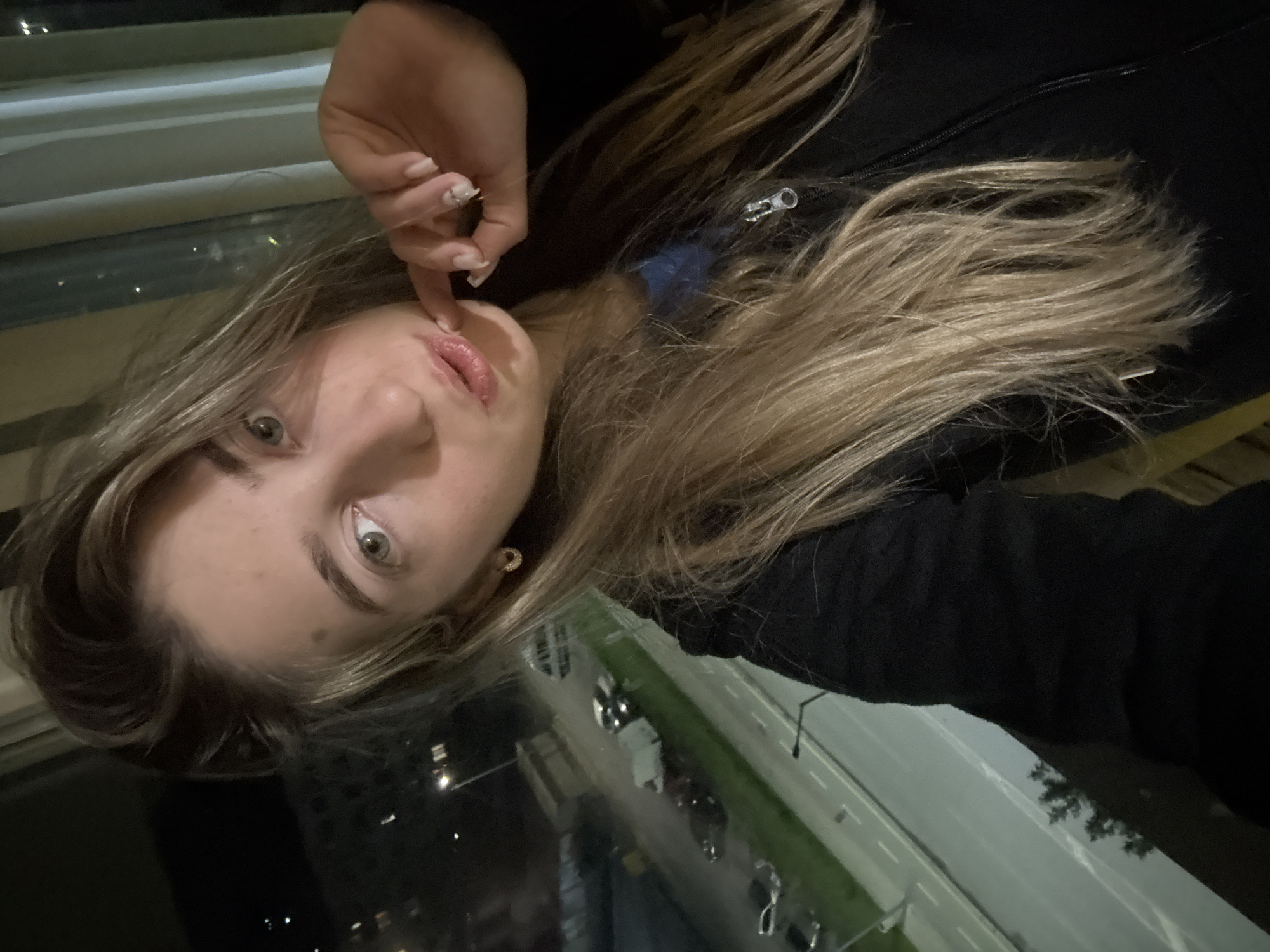 A woman with blond hair is posing for a selfie. She is wearing a black jacket, and her fingernails are painted white. She is touching her lips with her finger. The background shows a city at night, with streets, buildings, and trees visible through a window. Cars are visible on the street. The location appears to be elevated, possibly in a high-rise building.