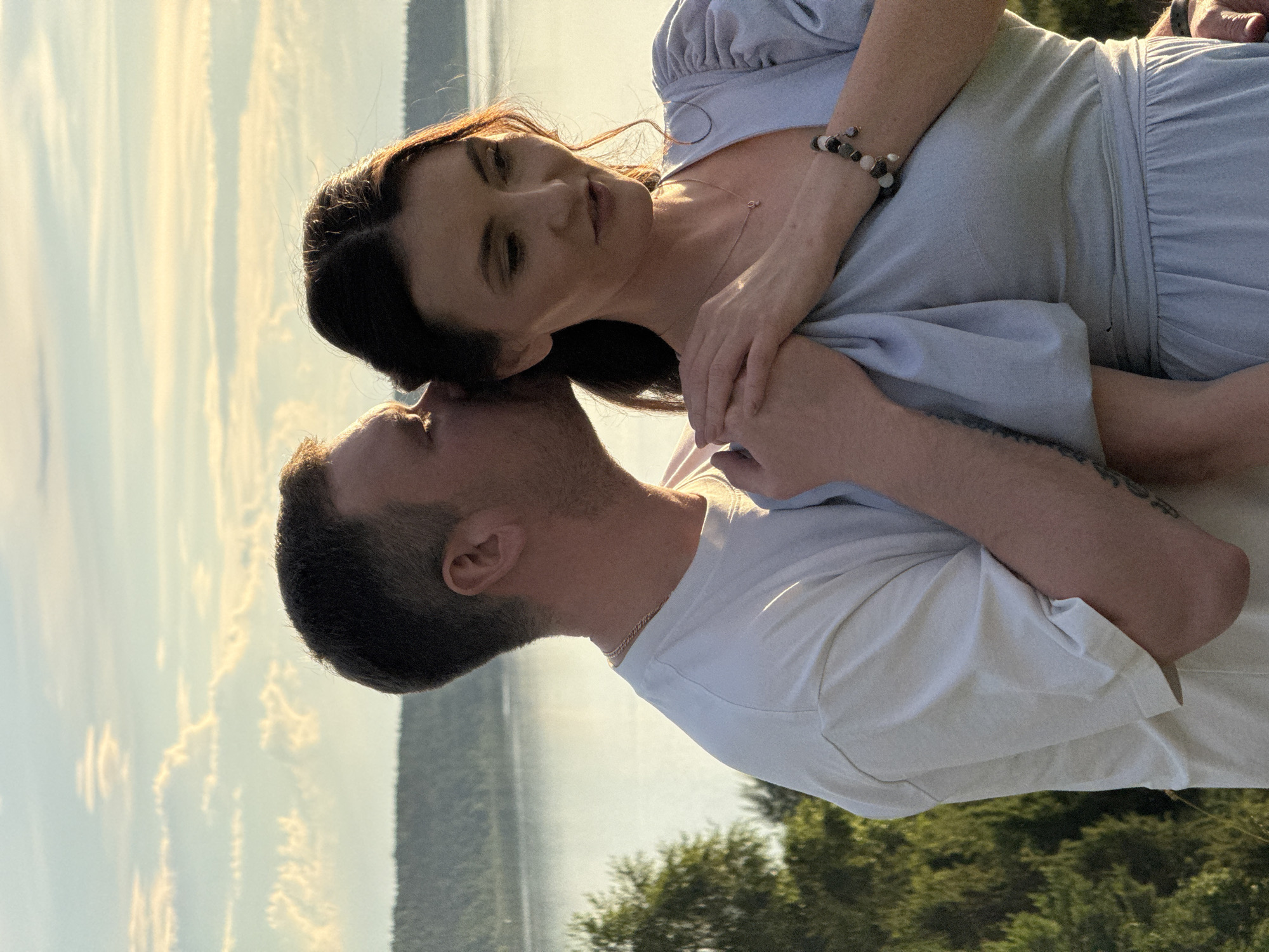 In Ольховка, Russia, a man is kissing a woman on the cheek. They are standing outdoors with a body of water and a tree line in the background. The man is wearing a white t-shirt and the woman is wearing a light blue dress. The sky above is filled with clouds.
