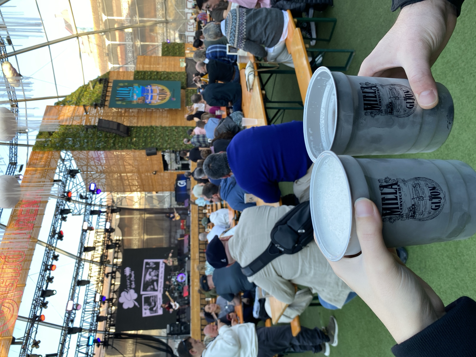 The image shows a view from a beer garden in Gijón, Spain. Two hands are holding large steins of beer, with the "Milla Cerveza de Gijón" logo visible on the steins. The beer is light-colored and topped with a thick head. In the background, there is a large gathering of people sitting at long wooden tables, with a stage set up for a band or performance visible in the distance. The environment appears to be an outdoor venue with a tent-like structure overhead, possibly for a festival or event. The atmosphere seems lively and festive.