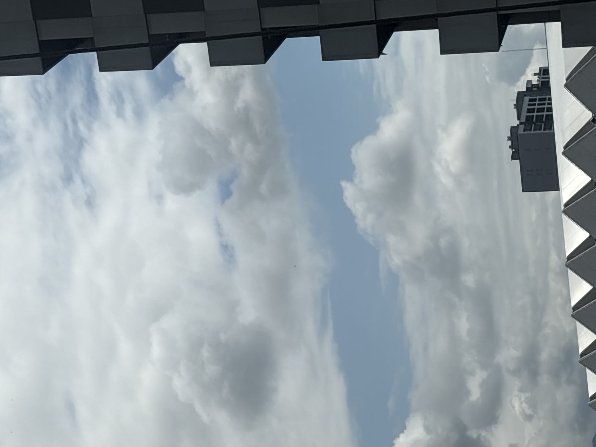 This media file is a photograph taken from a low angle, looking upwards. The scene is dominated by a cloudy sky, with patches of blue visible in between. The clouds are varied, with some appearing fluffy and white, while others are darker and more diffused.
Framing the sky are architectural elements. Along the top edge, there is a dark, metallic structure with a repeating geometric pattern, suggestive of a modern building's facade or overhang. On the right side, a portion of a white building with a distinctive, angled, layered design is visible. This design resembles a series of stepped triangular shapes.
Reflected within the reflective surfaces of these architectural elements, it is possible to see parts of buildings. On the right side, a dark, multi-story building with a grid-like pattern of windows is reflected. On the left, a similar, less distinct reflection of architectural features can be observed. The overall impression is that of an urban environment, possibly a city, where modern architecture interacts with the sky.
The lighting suggests it is daytime, likely mid-morning or afternoon, given the brightness of the sky and the contrast in the clouds. The weather appears to be partly cloudy. There are no people or animals visible in the frame. The image conveys a sense of looking up and out, perhaps from a ground-level or lower-story vantage point within a built environment. The mood is neutral and observational, focusing on the interplay of man-made structures and the natural sky. No text is visible in the image.