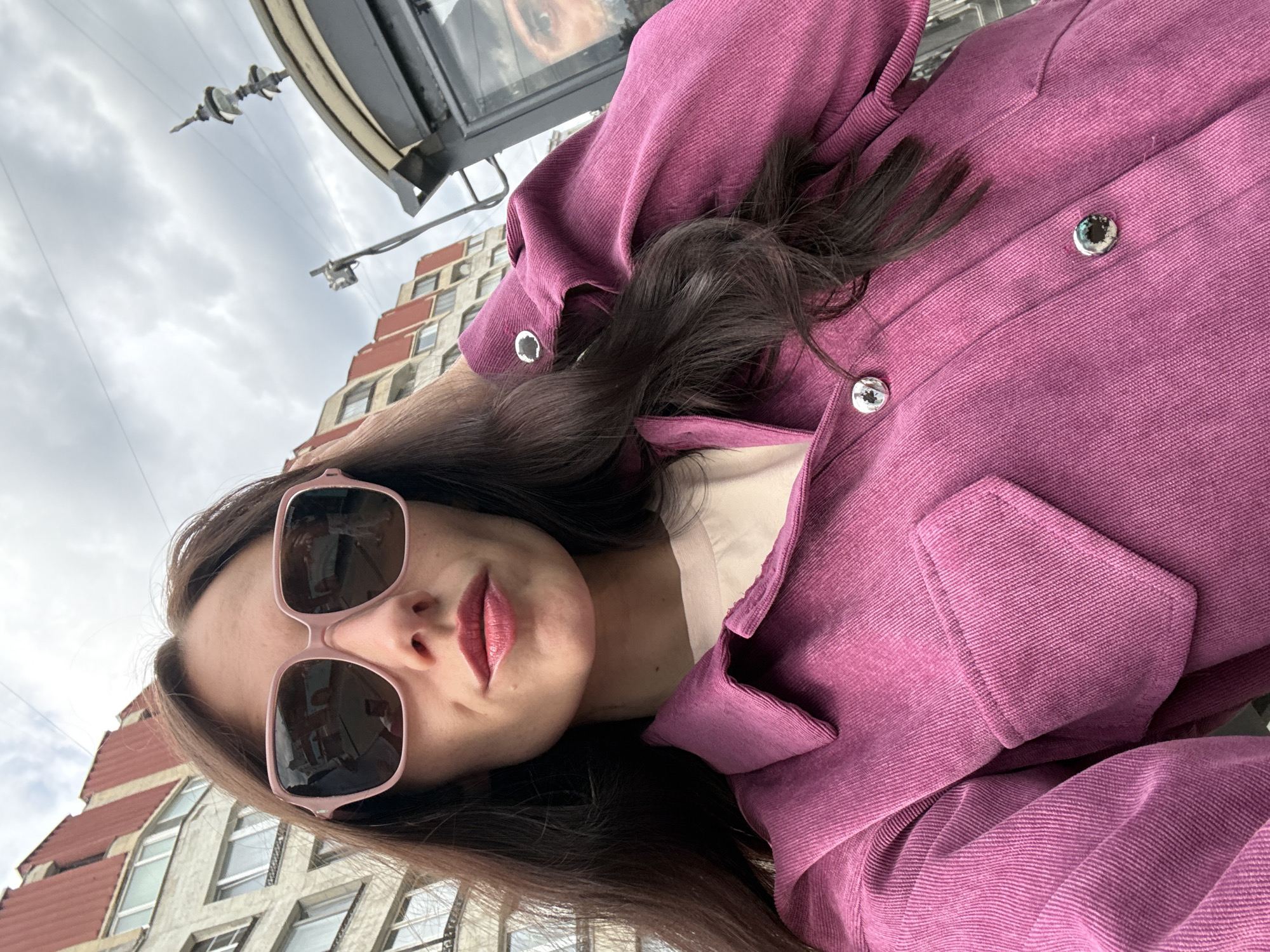 A low-angle, tilted selfie captures a woman outdoors in Saint Petersburg, Russia, against an overcast sky and urban backdrop. The woman, positioned in the lower left of the frame, looks slightly away from the camera. She has long, dark hair, and her eyes are obscured by large, light pink-framed sunglasses. Her lips are painted with pink lipstick.
She wears a vibrant purple or fuchsia jacket, which appears to be made of corduroy due to its distinct ribbed texture. The jacket features large, round silver buttons down the front and silver eyelets on the cuffs. A light-colored top is visible underneath her jacket.
In the background, a building with distinctive reddish-brown sloped roof sections and multiple windows suggests an older European architectural style. Above the woman, overhead wires, likely for public transport like trams, stretch across the sky. Dark, metallic structures are also visible, possibly part of a bus stop or other street furniture. Notably, a blurry reflection resembling a dog's face can be seen in a dark, reflective surface in the upper background, possibly a window or screen on a public transport shelter. The cloudy sky indicates a non-sunny day. No discernible text is visible.