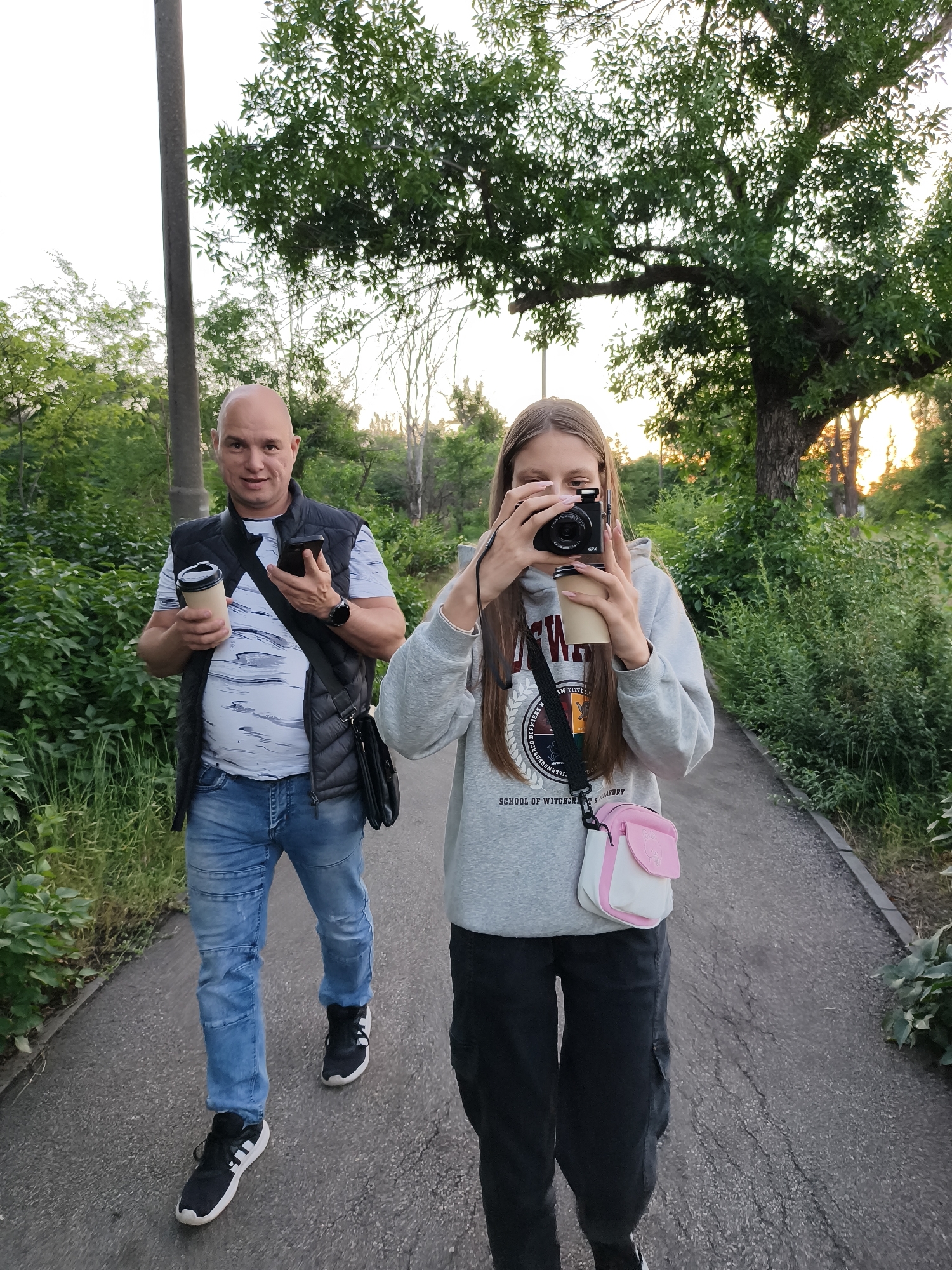 Two individuals, a man and a teenage girl, are walking along a paved path in an outdoor, green environment, likely a park or suburban area. The man, on the left, is bald, wearing a grey t-shirt with a dark vest over it, blue jeans, and black and white athletic shoes. He is holding a disposable coffee cup in his left hand and a smartphone in his right hand, with a smartwatch on his left wrist. A dark shoulder bag is slung across his chest. The girl, on the right, has long brown hair and is wearing a grey hooded sweatshirt with a graphic design and text that reads "SCHOOL OF WITCHCRAFT & WIZARDRY," black cargo pants, and black athletic shoes. She is holding a digital camera to her face, appearing to take a photograph, and is also holding a disposable coffee cup. A small, pink and white crossbody bag is visible on her front. The path is bordered by lush green vegetation and trees. The lighting suggests it is late afternoon or early evening. The scene is captured from a low angle, looking slightly upward. It is not possible to determine the exact location within Kryvyi Rih, Ukraine from the visual information provided.