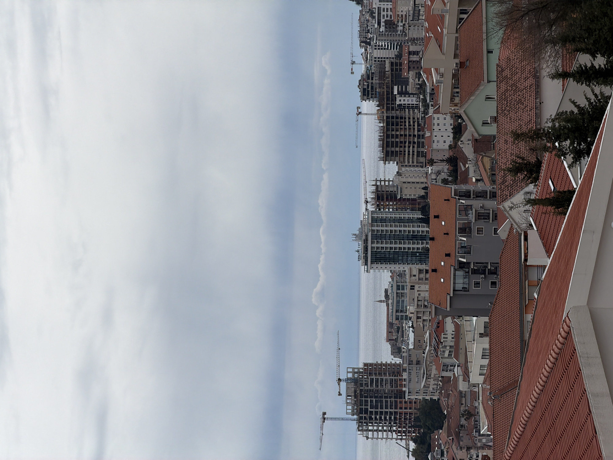 The image shows a cityscape, likely Budva, Montenegro, as evidenced by the building styles, proximity to water, and ongoing construction. The photo is taken from an elevated position, looking down at buildings with red tile roofs, interspersed with modern high-rises under construction. Several cranes are visible against the backdrop of the sea and cloudy sky. The buildings are a mix of residential and commercial structures. The sky is overcast with a uniform distribution of clouds.
