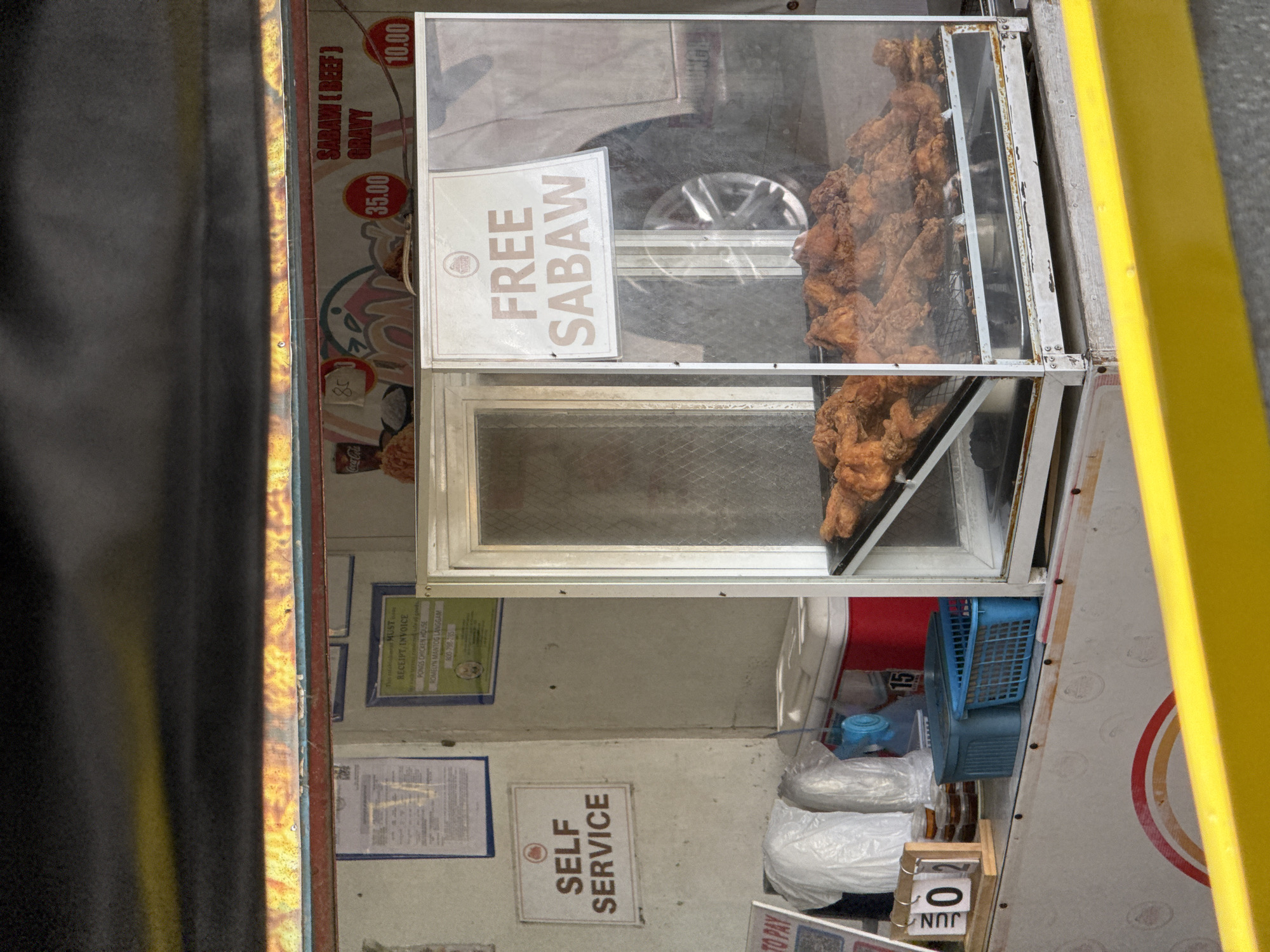 This image captures a vibrant street food stall, likely mobile or roadside, specializing in fried chicken. The central feature is a glass display case filled with numerous pieces of golden-brown, crispy deep-fried chicken, stacked invitingly. A prominent white sign inside the display proclaims "FREE SABAW" (free broth).
The stall operates on a "SELF SERVICE" model, as indicated by another sign. Various items are displayed or stored within the stall, including a blue plastic basket, white plastic bags, and a small blue container. A menu or price list board is partially visible, showing "SABAW (BEEF) 35.00" and "GRAVY 10.00," alongside other prices like "10.00" and "85" near a "Coca-Cola" logo.
Key location cues visible include a framed "MUST RECEIPT INVOICE" with the business name "PONGS CHICKEN HOUSE" and contact "ROMELYN MANTOG LANGGAM," along with a phone number "800-798-285." This information, coupled with the term "Sabaw," firmly places the scene in the Philippines, consistent with the Davao City context. The reflection of a car wheel in the display glass suggests a busy, possibly urban, outdoor setting during daylight hours. A small wooden stand with "JUN" and a "0" is also present, possibly indicating a date or numbering. The overall impression is that of a bustling, local eatery offering affordable and accessible food.
