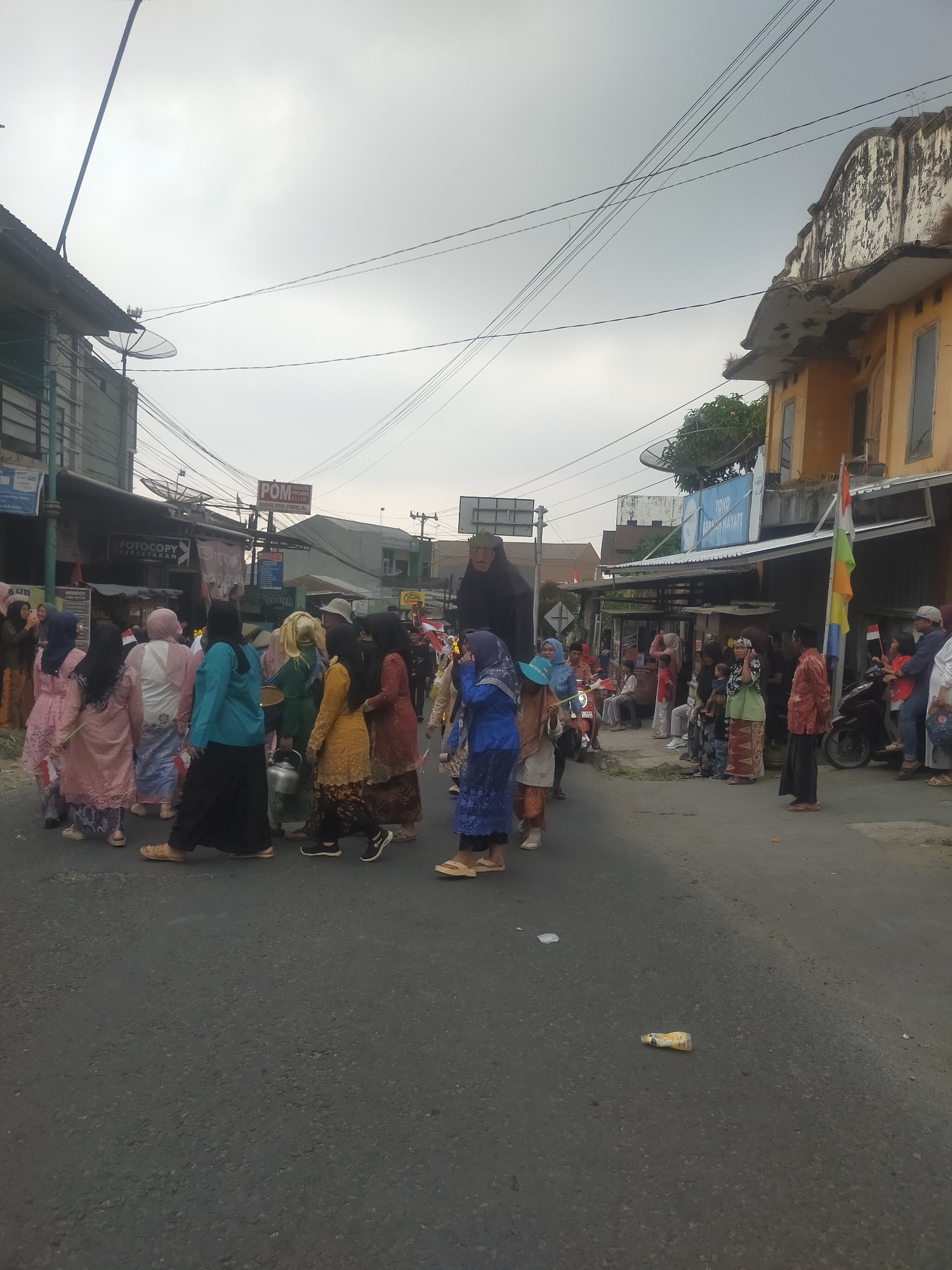 This is a photograph taken in Tuwel, Indonesia, showing a procession on a street with buildings on either side. The sky is overcast and gray. On the left side of the street are several buildings, one with a satellite dish on its roof and another with a sign that reads "FOTOCOPY PERCETAKAN". On the right side of the street, there are more buildings, one of which is yellow with a weathered facade. A tall, dark figure, possibly a puppet or costume, is prominently visible in the procession, being carried or walked by people. The procession consists of a group of people, many of whom are wearing traditional Indonesian attire, including long dresses and headscarves. Some individuals are carrying small Indonesian flags. There are also motorcycles and a few spectators on the side of the road. The street is paved with asphalt and shows signs of litter, including a discarded bottle. The overall scene depicts a cultural event or celebration taking place in a semi-urban environment.