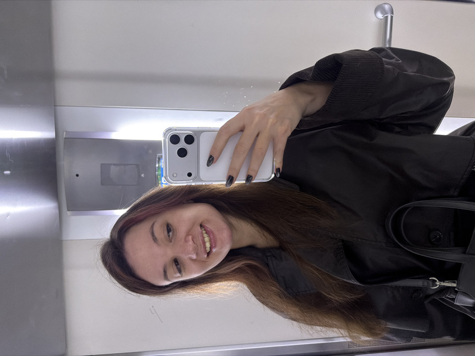 A person with long brown hair smiles, holding up a white smartphone with a clear case. They are wearing a dark jacket with a ribbed cuff. A black bag hangs from their shoulder. The background is a mirrored surface of an elevator, reflecting the light. A metal handrail is visible above. A grey control panel with a button is also reflected in the elevator mirror.