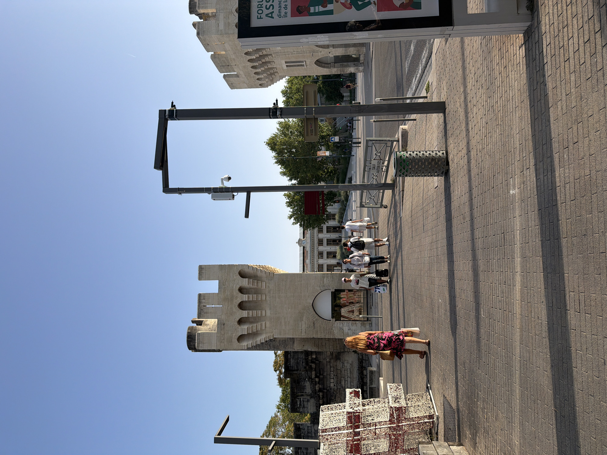 The image depicts a street scene in Avignon, France, likely near a historic fortified structure. In the foreground, a brick-paved sidewalk stretches across the frame, illuminated by the warm light of late afternoon, casting long shadows. A woman in a floral dress walks away from the camera, carrying shopping bags and a straw purse. To her left, a large, decorative, cube-shaped structure composed of white and red wire frames stands near the edge of the sidewalk.
Dominating the mid-ground is a section of ancient stone fortification with crenellations and arched openings, appearing to be part of a tower or wall. A modern doorway with a glass entrance is visible within one of these arches, with "A" shapes displayed inside. To the right of this structure, another section of similar stone fortification is visible, continuing along a street.
Several people are walking on the sidewalk and crossing the street. Some are dressed casually in t-shirts and shorts, carrying shopping bags or backpacks. Streetlights and security cameras are mounted on tall poles along the sidewalk. In the background, a building with a clock tower is visible, suggesting a public square or transportation hub. The sky is clear and blue. A large advertisement panel stands on the right side of the frame.