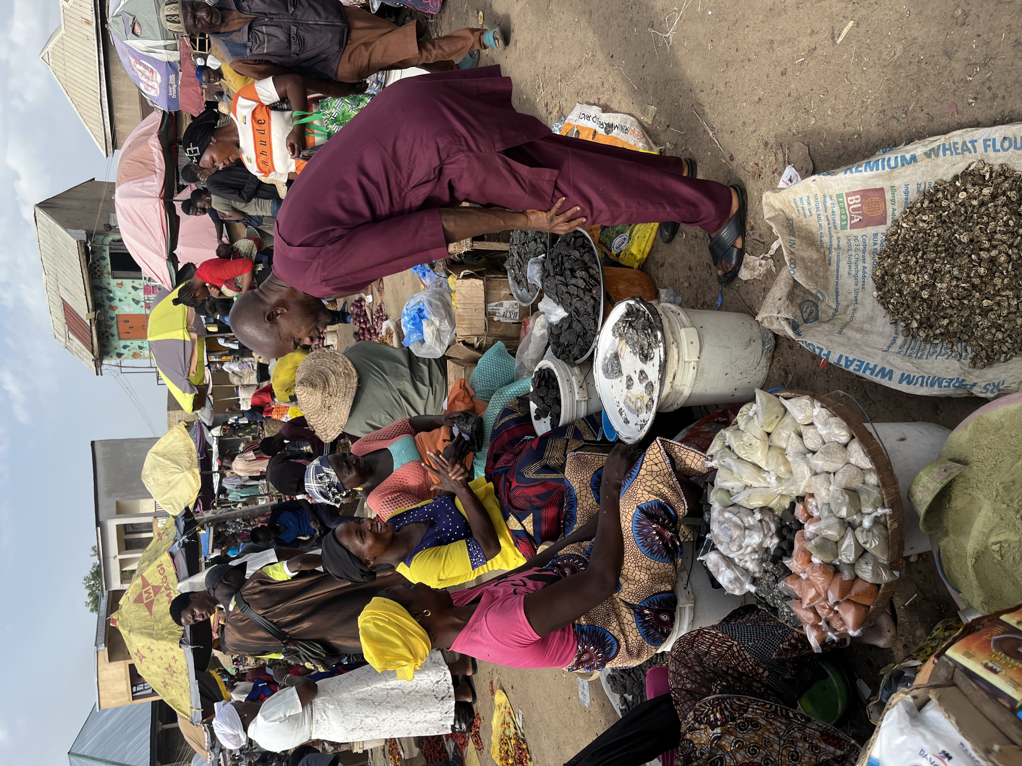 The image depicts a bustling outdoor market scene in Rafin Par, Nigeria. In the foreground, a man in a maroon traditional outfit is bent over, interacting with a stall. The stall displays an assortment of goods, including small, dark dried items in metal bowls, and various spices and powders packaged in plastic bags and placed within a woven basket. To the left, a woman in a pink shirt and a colorful patterned skirt sits next to a similar assortment of packaged goods, while another woman in a yellow shirt and a patterned skirt is visible behind her. A third woman, wearing an orange top and a patterned skirt, is seated further back.
Behind the stall, various market activities are visible. People mill about, some under the shade of colorful umbrellas. Buildings with tin roofs and textured walls are present in the background. The ground is unpaved, consistent with an outdoor market environment. The overall impression is one of a vibrant and active marketplace.