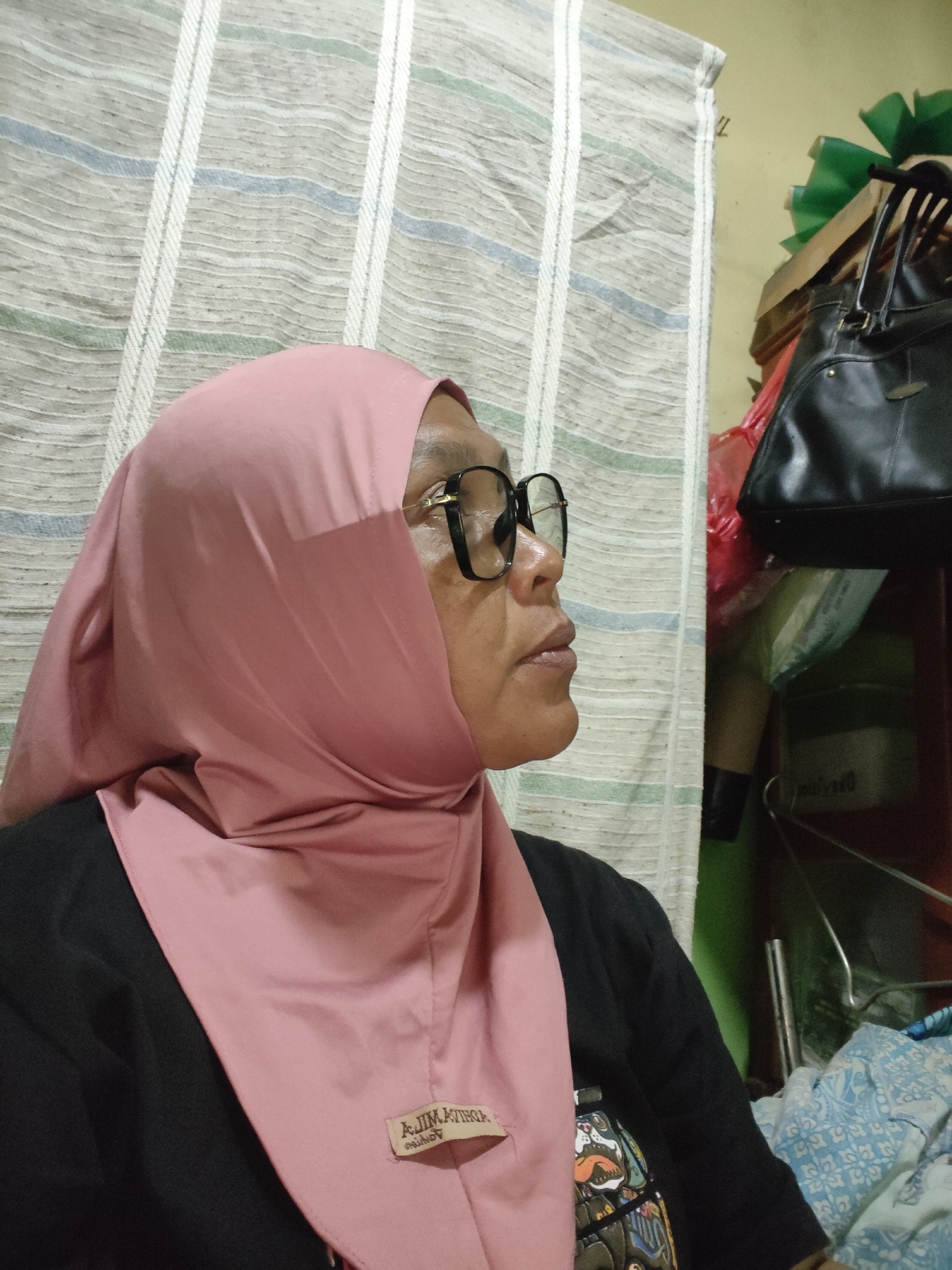A middle-aged person with glasses, wearing a pink hijab and a black t-shirt, is shown in a profile view against a striped curtain. The person's head is turned to the right, and they appear to be looking away. In the background, a black handbag is visible on a shelf, along with other items. The setting appears to be indoors, possibly a bedroom or a living area in a residential building located in Lori, Indonesia.