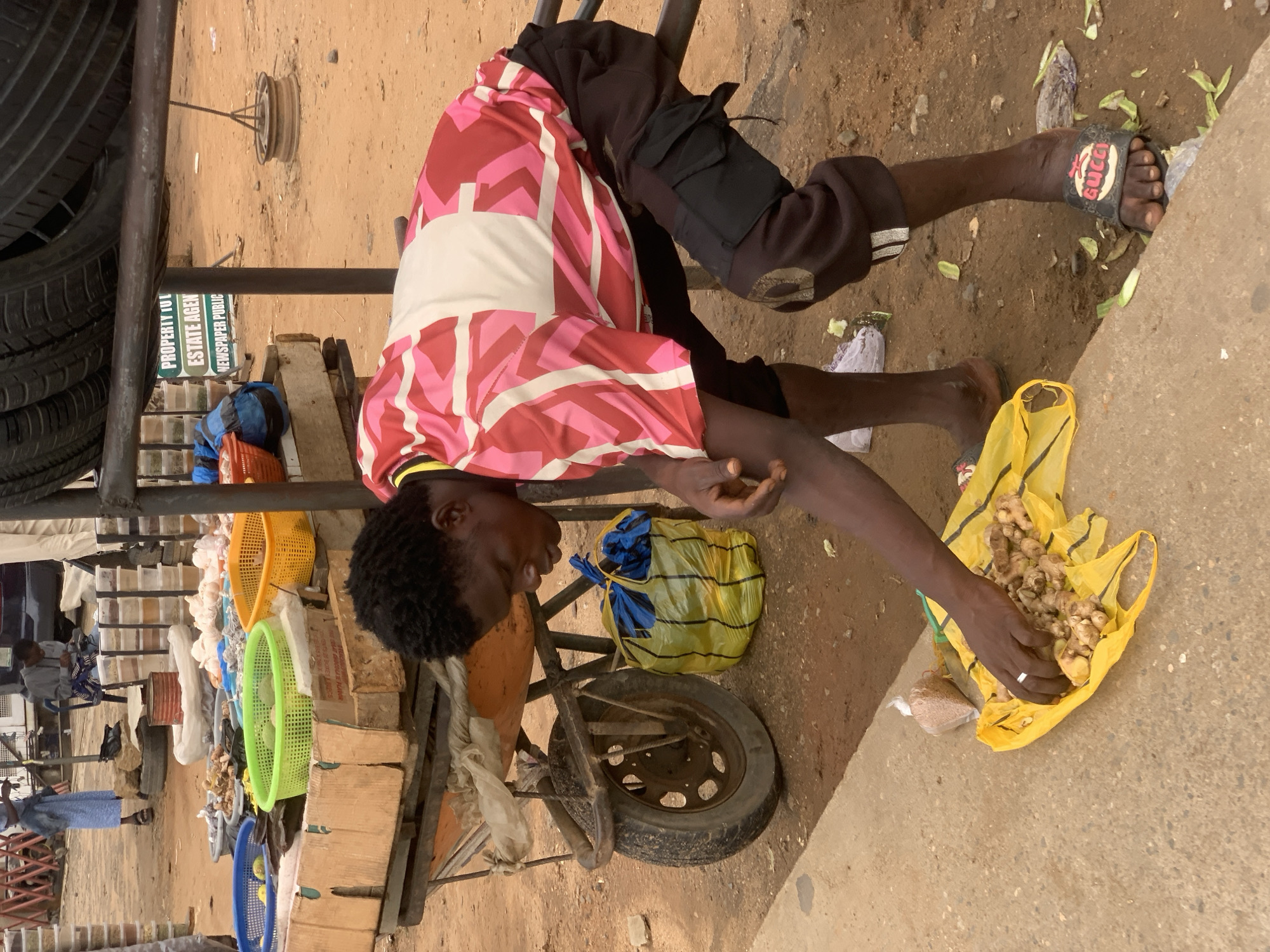 An individual sits on a makeshift seat with a cart in Ikorodu, Nigeria. He wears a red and pink patterned shirt, dark-colored shorts, and sandals with a Gucci logo. He is sorting through ginger in a yellow plastic bag. The cart contains items such as baskets, produce, and tires. A sign for a real estate agent is visible in the background. The environment is dusty with evidence of discarded food or vegetable matter on the ground.