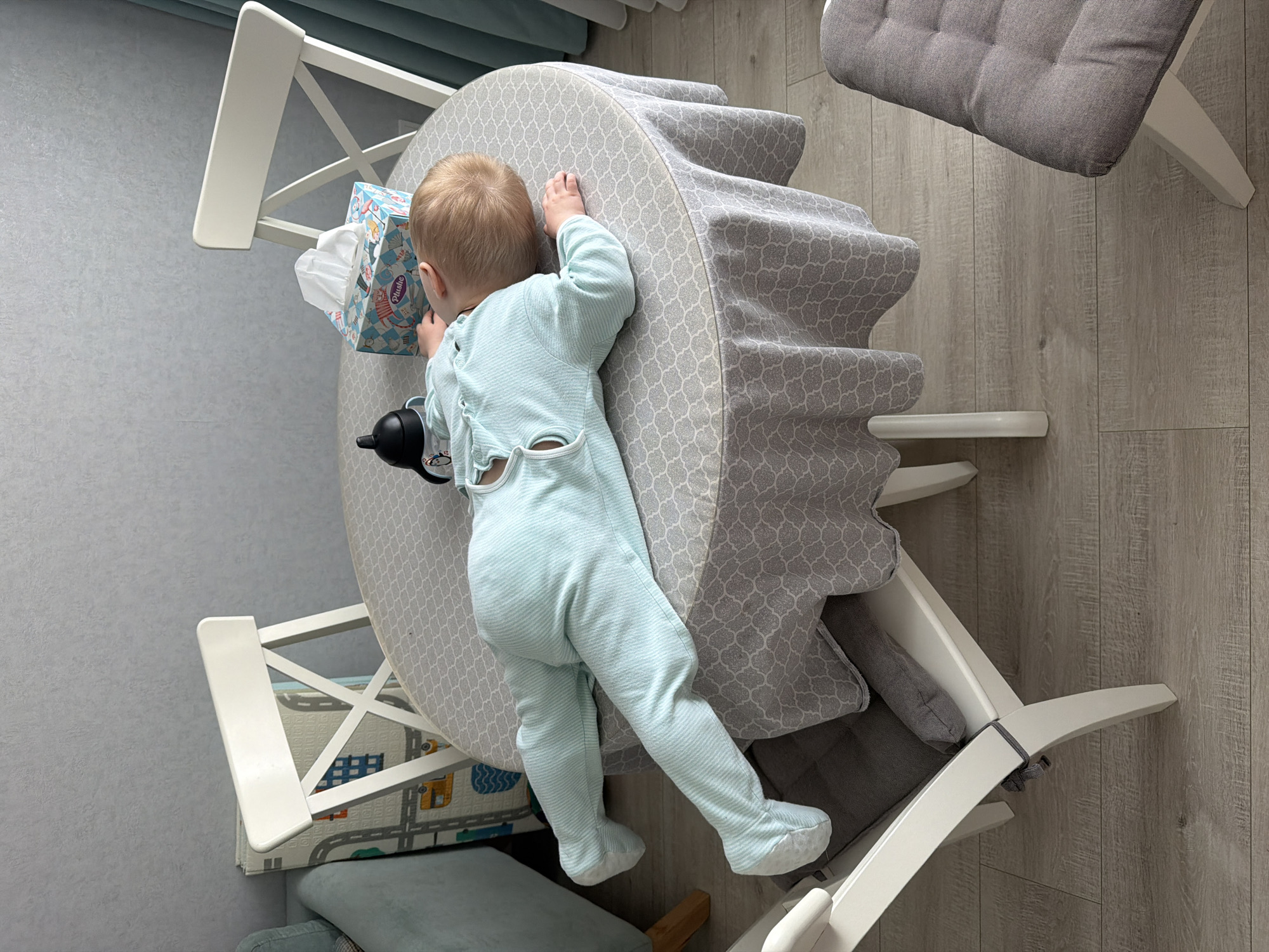 The scene, located in Votkinsk, Russia, depicts an indoor room, likely a dining area or kitchen. A young child, appearing to be an infant or toddler with light blonde hair, is lying face down on a round table. The child is dressed in a light blue, footed sleepsuit with a subtle striped texture and visible snap fasteners along the back. The table is covered with a light gray tablecloth featuring a repeating white quatrefoil pattern. On the table, to the child's right, rests a light blue and white "Plusho" brand tissue box, and to the child's left, a black baby bottle or sippy cup with a white graphic design. Three white wooden chairs with X-shaped backrests and gray upholstered seat cushions are visible around the table. The room features light gray walls, light wood-look laminate flooring, a light blue curtain partially visible on the right, and a children's playmat with a road-and-city design leaning against a wall in the background on the left.