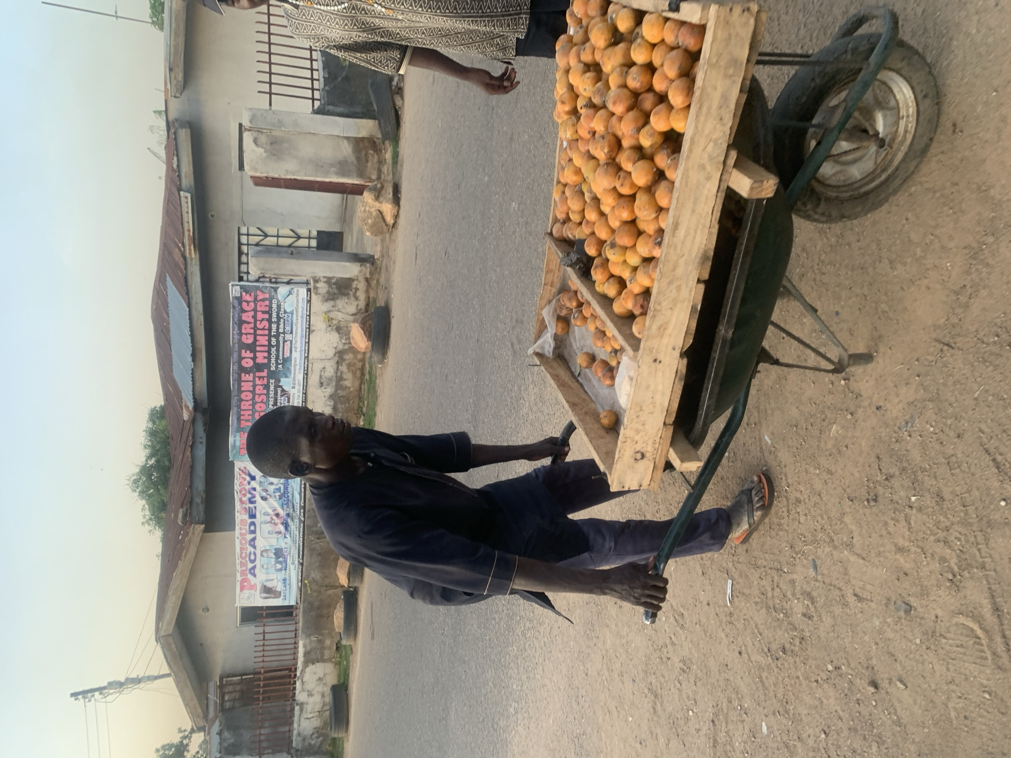 The image depicts a vibrant street scene in Jos, Nigeria, featuring a man pushing a wheelbarrow filled with fruit. The central figure, a man wearing a dark blue long-sleeved shirt, dark pants, and sandals, is actively pushing a green wheelbarrow modified with a wooden crate on top. This crate is overflowing with a multitude of small, round, orange-colored fruits, likely African star apples (agbalumo) or a similar local variety, suggesting he is a street vendor. Another person, partially visible on the right, stands near the wheelbarrow, wearing a distinctive patterned shirt.
The setting is an urban environment with a combination of paved road and dirt shoulder. In the background, various buildings are visible, including a single-story structure with a rusty corrugated metal roof and concrete walls, characteristic of local architecture. Utility poles and repurposed tires serving as roadside barriers add to the authentic street atmosphere. The clear, bright sky indicates it is daytime, possibly late afternoon or early morning.
Prominently displayed on a building in the background are two banners. One advertises "THRONE OF GRACE GOSPEL MINISTRY," detailing a "SCHOOL OF THE SWORD (A Community Bible Class)" held "THUR 4:30PM," along with contact information. The other sign promotes "PRECIOUS STONE ACADEMY," offering "DAYCARE NURSERY PRIMARY SECONDARY" education with the slogan "Every Child A Gem; Building a strong foundation for tomorrow." The image captures an ordinary moment of daily commerce and life in a Nigerian city.