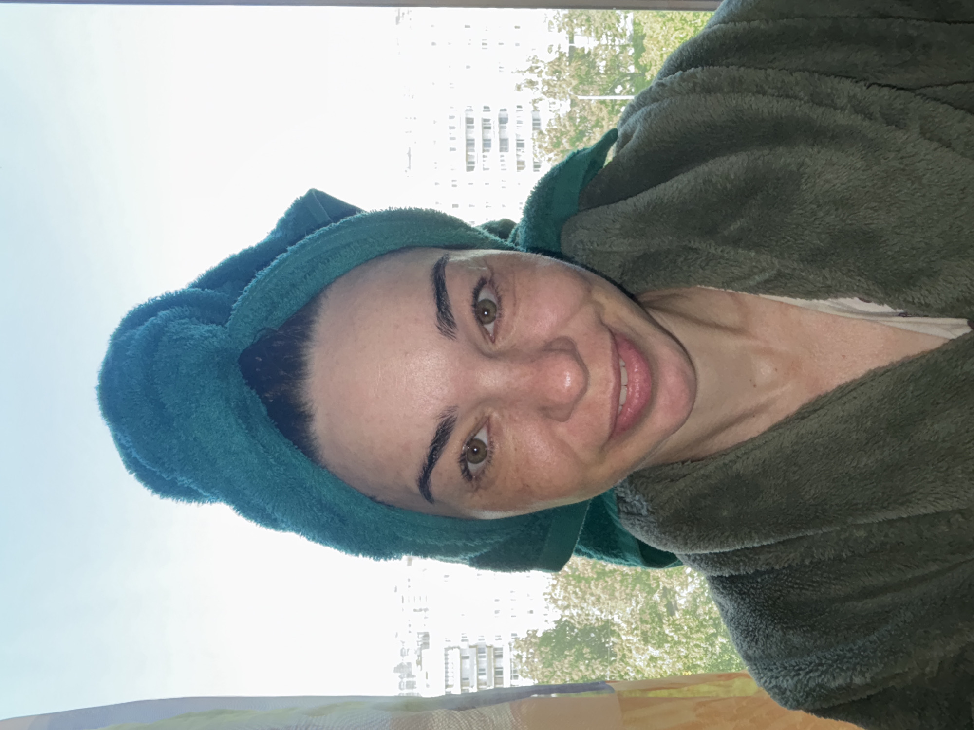 A medium close-up shot, rotated 90 degrees counter-clockwise, features a person, likely female, smiling directly at the camera. Her head is wrapped in a teal-colored terrycloth towel, suggesting she may have recently washed her hair. She is wearing a dark green, fuzzy robe. Her facial features include dark eyebrows, prominent green eyes, and visible freckles across her nose and cheeks. Her lips are slightly parted in a cheerful smile, revealing a hint of teeth.
The background shows a bright, sunlit outdoor scene viewed through a window. A clear, light blue sky fills the upper portion of the background. Below the sky, blurred architectural structures, appearing to be multi-story buildings, are visible, along with green foliage from trees, indicating an urban or suburban setting. The strong natural light suggests it is daytime. The overall mood conveyed by the subject's expression is one of happiness or contentment. There is no visible text in the image.
