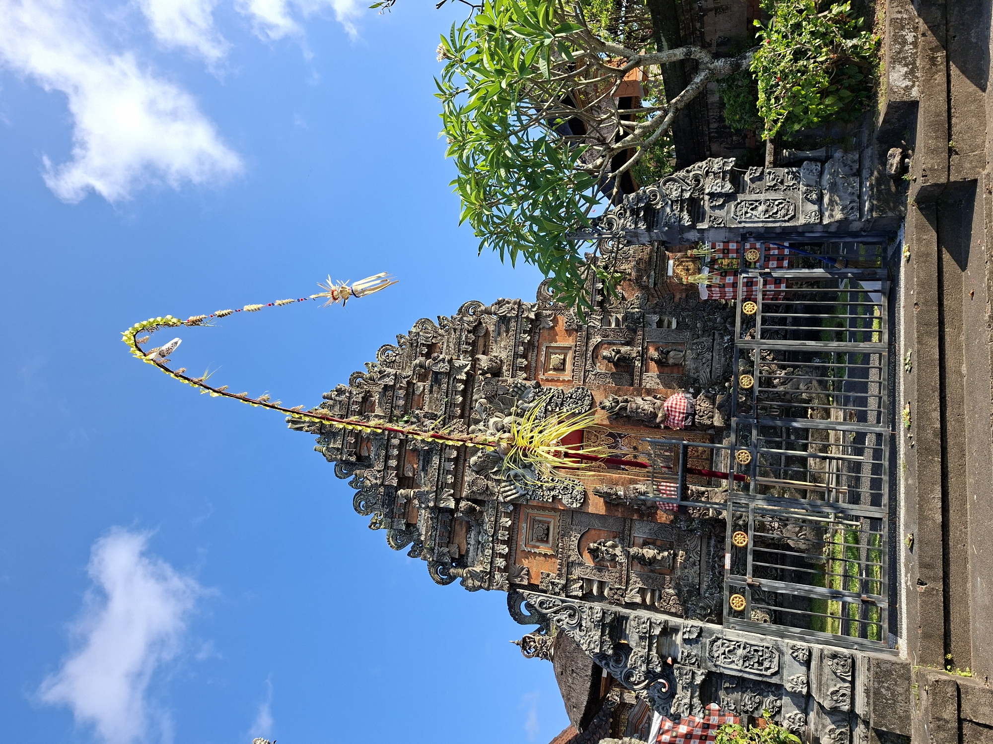 An ornate stone temple structure stands against a clear blue sky with a few wispy clouds. The temple features intricate carvings, sculptures, and tiered rooflines. A tall, curved banner adorned with colorful decorations and streamers extends from the top of the temple towards the sky. In front of the temple is a metal gate, and to the right, a lush green tree with white flowers is visible. The ground in front of the gate consists of stone steps. The scene appears to be a place of cultural or religious significance.